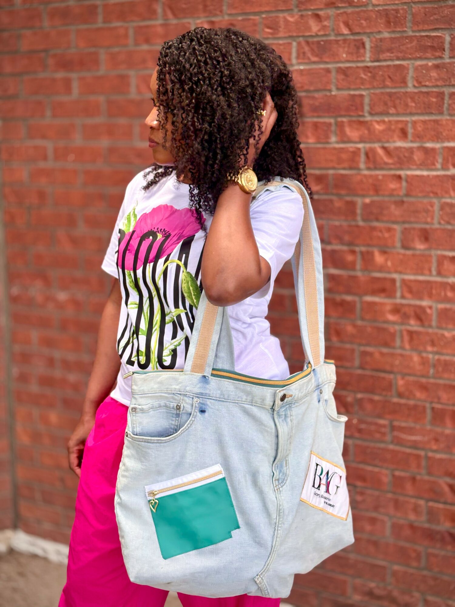 Person with curly hair wearing a white t-shirt and pink pants, carrying a large light blue bag with green and white pockets, standing against a brick wall.