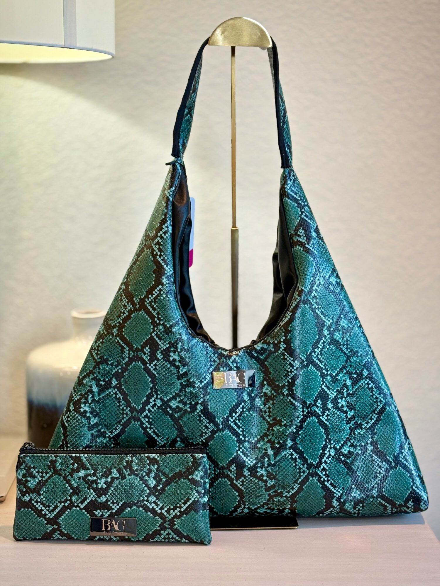 Large teal handbag with snake print pattern, matching smaller wallet, both on a table against a light wall.