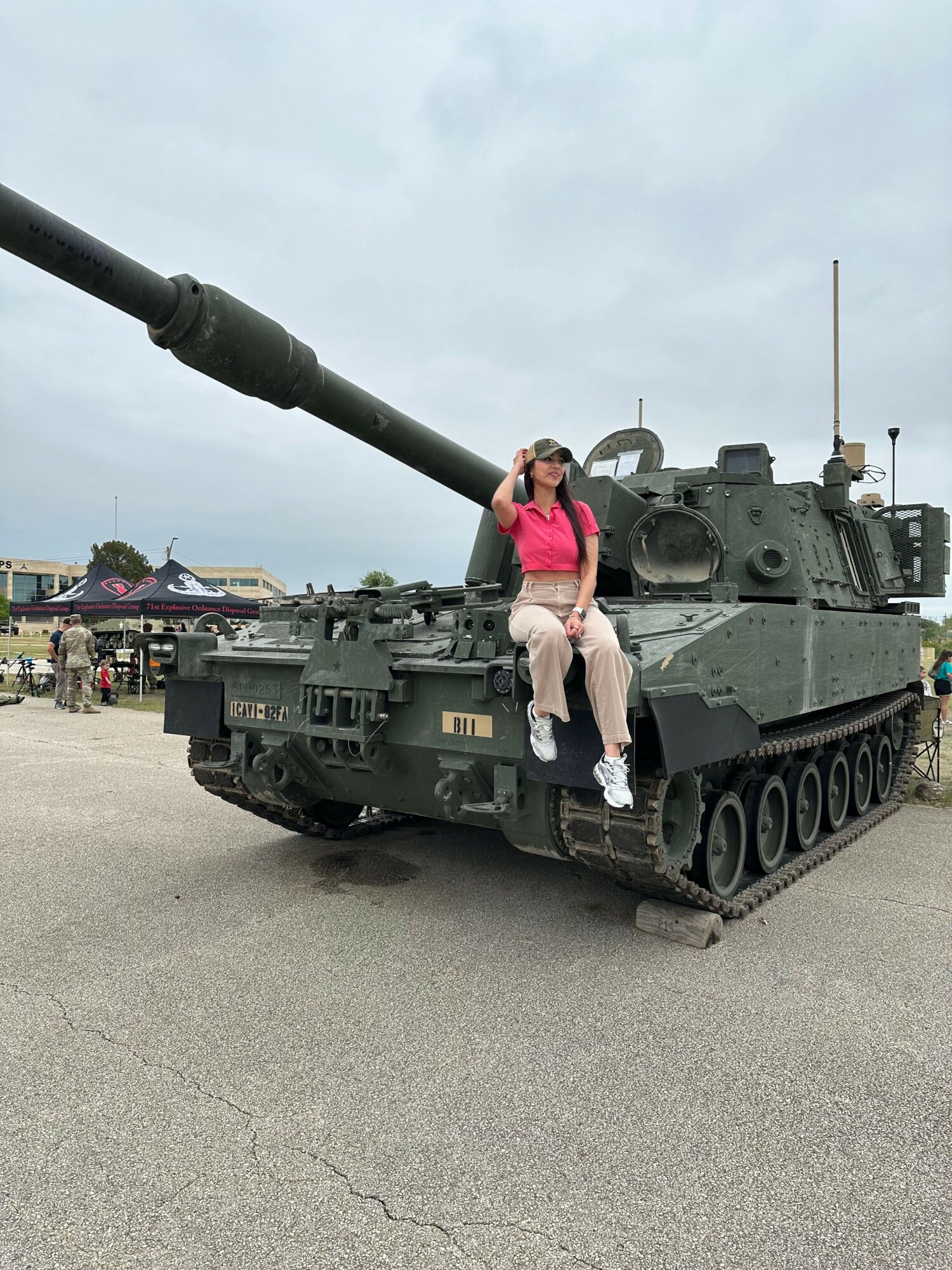 Person standing on a military tank with a large gun barrel, in an outdoor setting with buildings in the background.