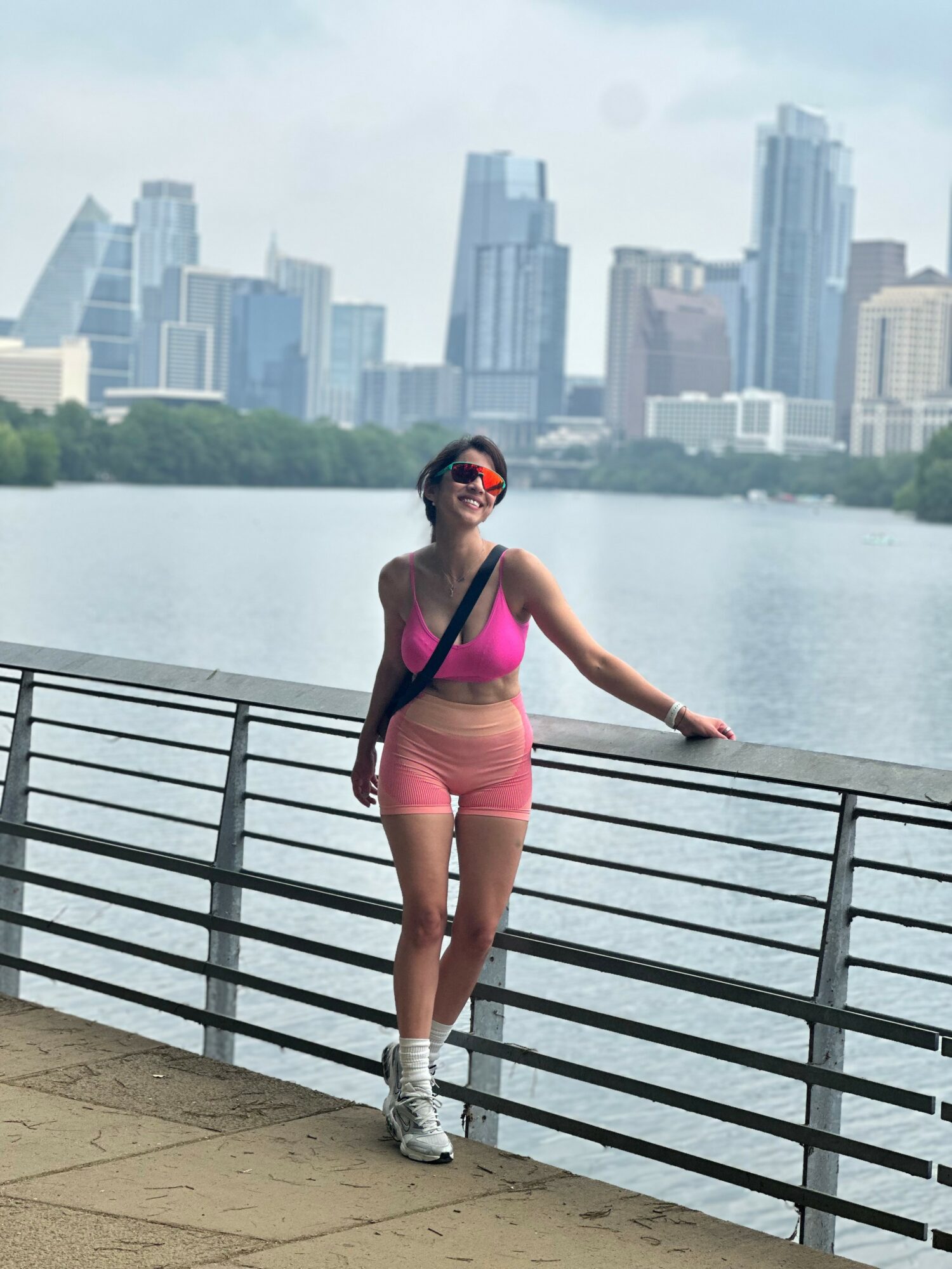 Woman in pink top and shorts leaning on a railing near water with city skyline in background.