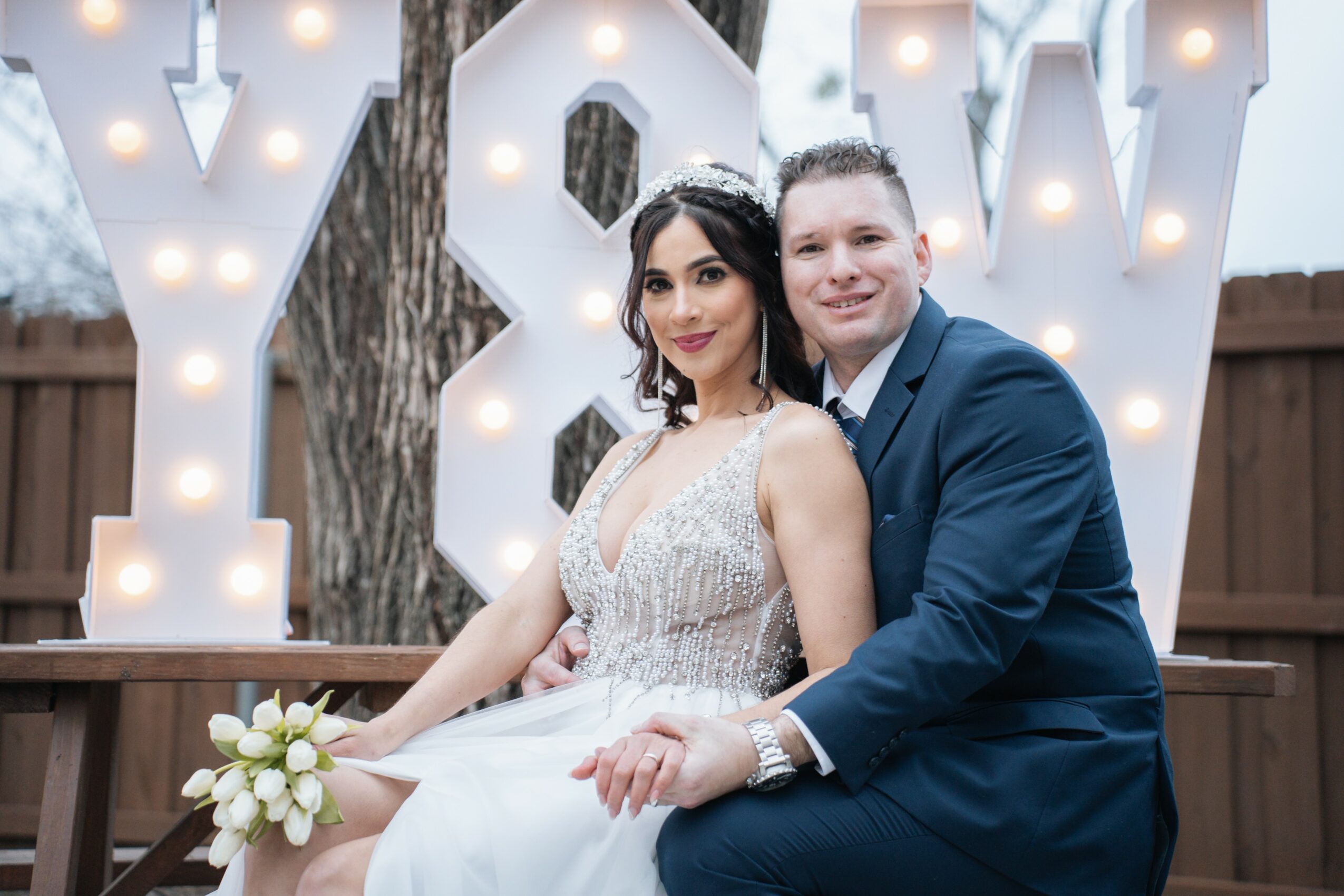 A bride and groom sit together outdoors in front of a large illuminated decorative sign, smiling at the camera.