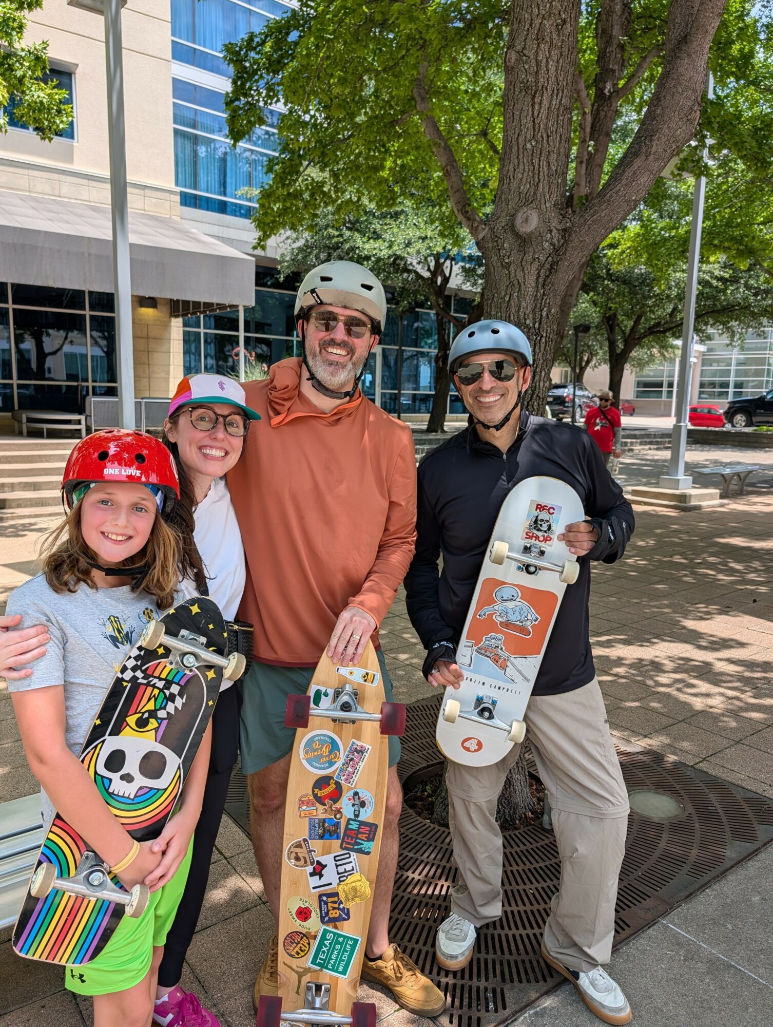 Four people with skateboards and helmets standing outdoors near a tree, smiling, in an urban area with buildings and a sidewalk.