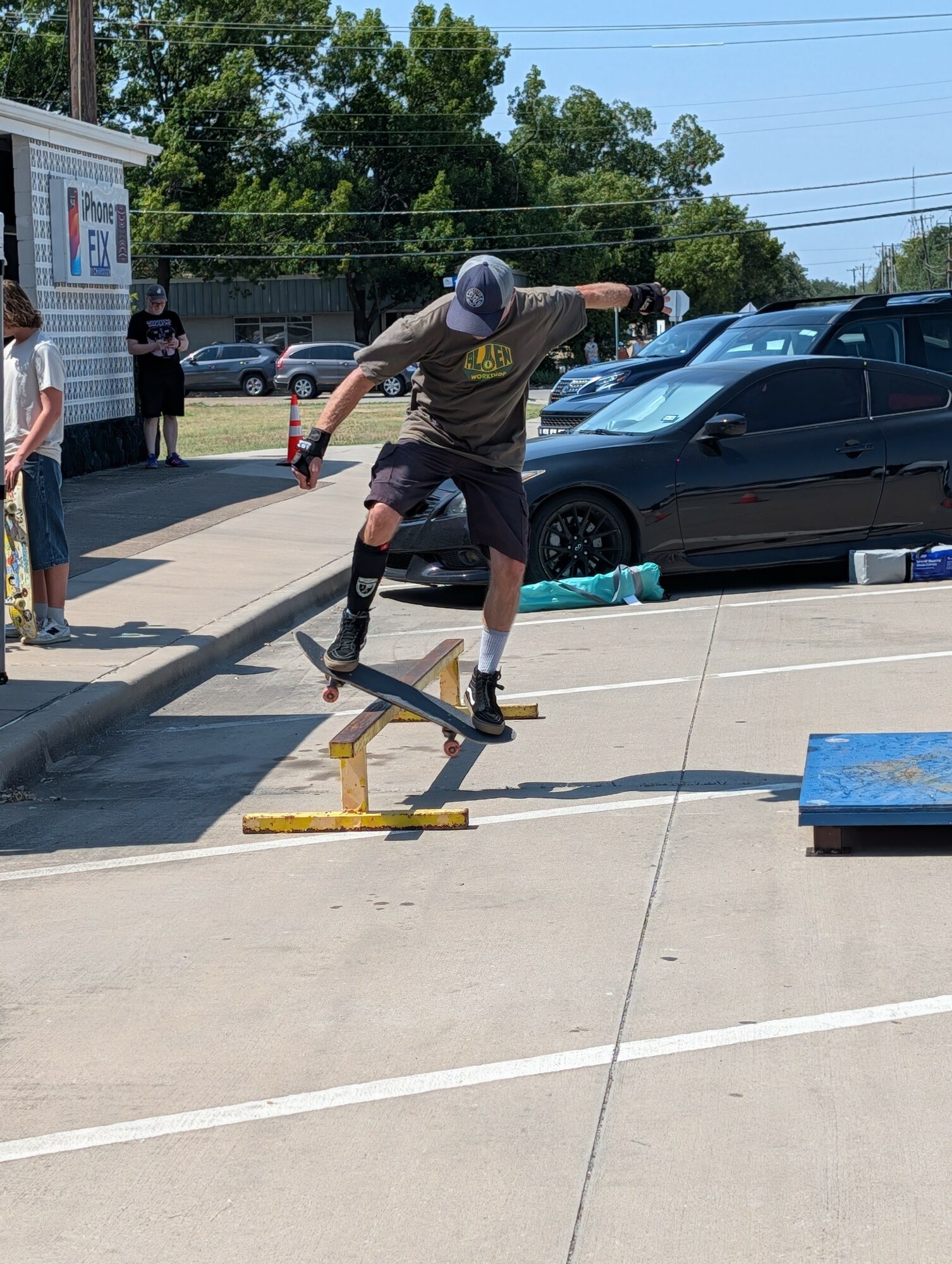 Skateboarder in black shorts and a gray shirt performs a trick on a rail in a parking lot, with onlookers nearby.