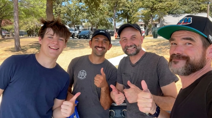 Four men smiling outdoors, giving thumbs-up, with trees and parked cars in background.