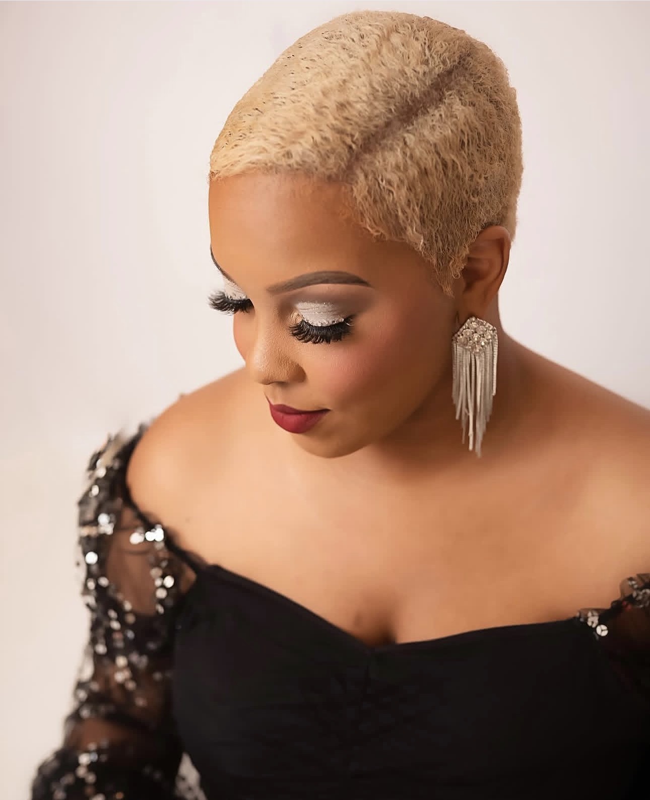 Woman with short blonde hair, makeup, large earrings, wearing a black dress with sheer, embellished sleeves, looking downward.