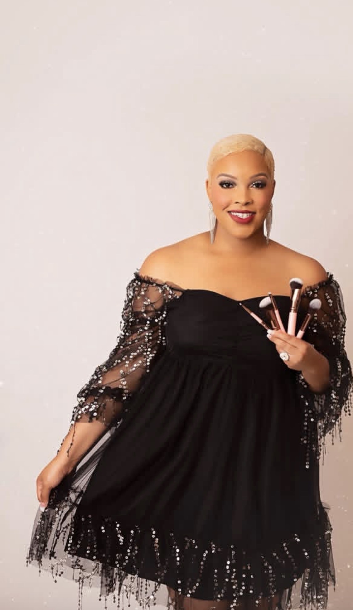 Woman with short blonde hair in a black off-shoulder dress with sheer, beaded sleeves, smiling and holding a makeup brush.