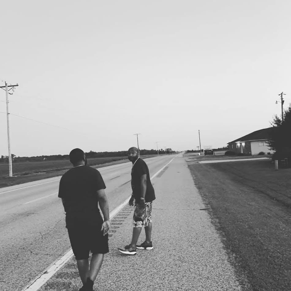 Two people walking on a road in a rural area, with power lines and a building in the background, in black and white.