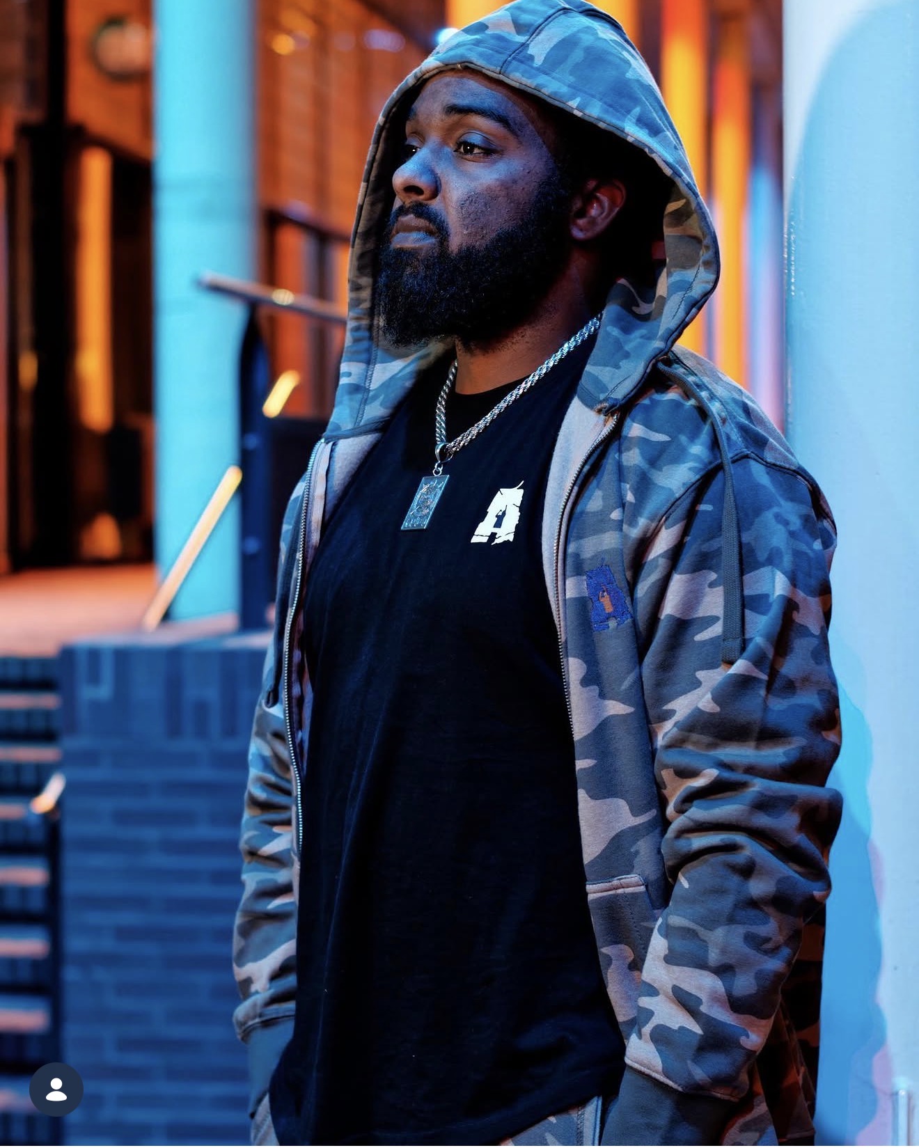 Man with a beard wearing a camouflage hoodie and black shirt, standing outdoors at night with colorful lights in background.