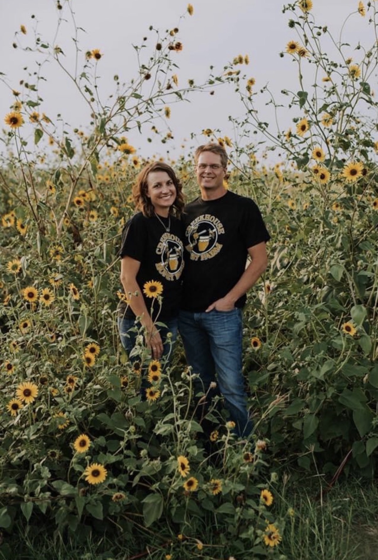 Two people standing in a sunflower field, smiling, with tall sunflowers around them, overcast sky in background.