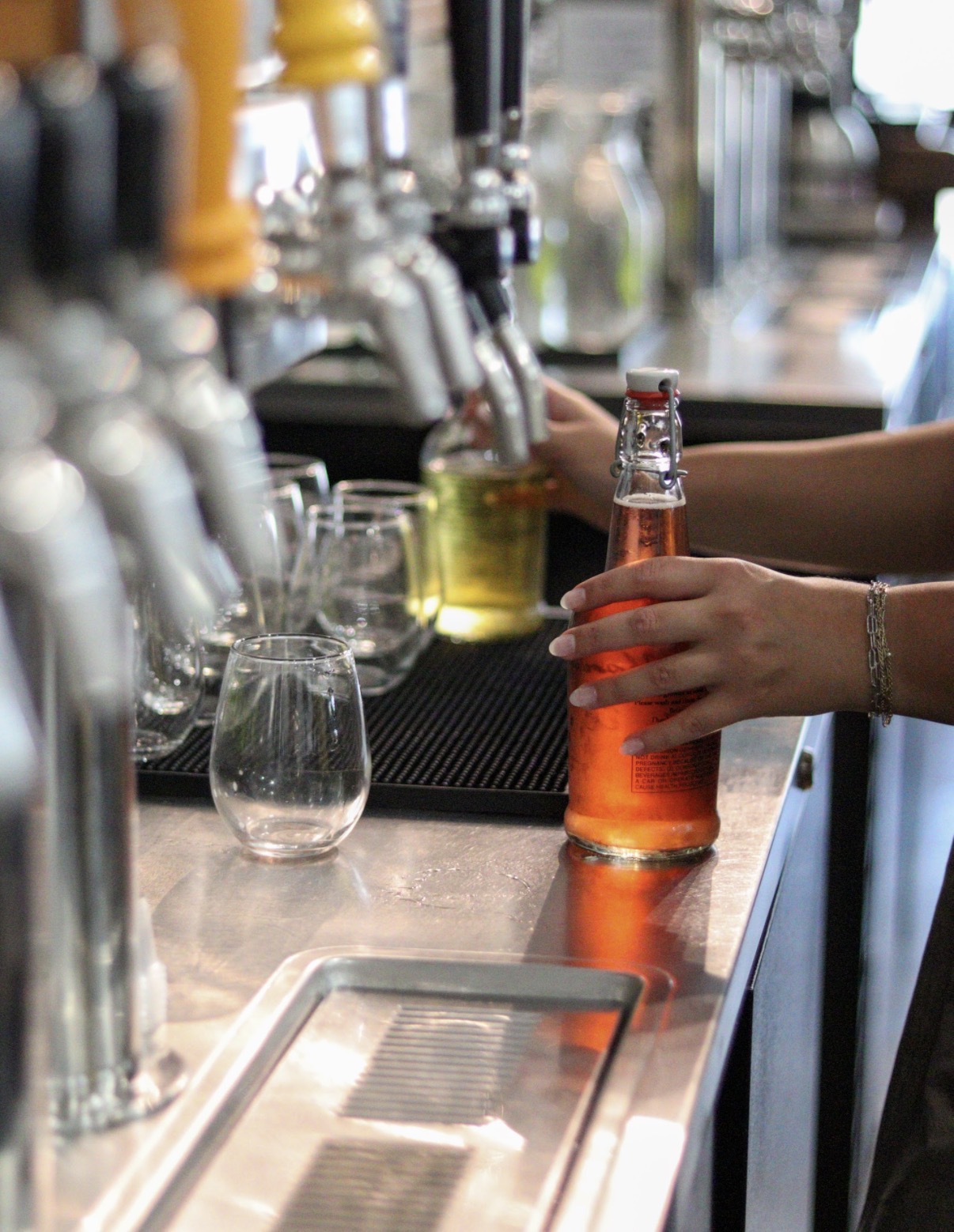 Person pouring orange-colored drink into a glass at a bar counter with taps and glasses nearby.