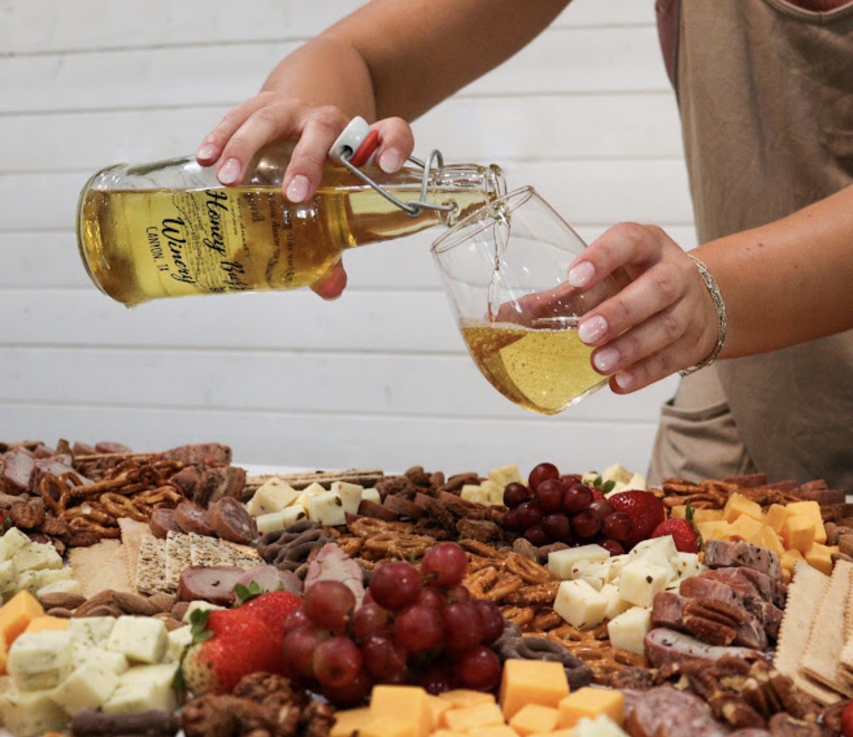 Person pouring yellow wine into a glass over a table with cheese, grapes, and snacks, against a white background.