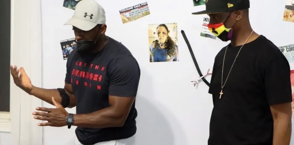 Two men standing in front of a wall with photos and posters, one wearing a white cap and the other a colorful mask.