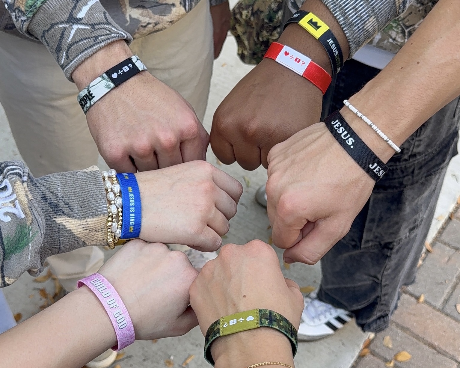 Five people show fists together, wearing various wristbands and bracelets, in an outdoor setting.