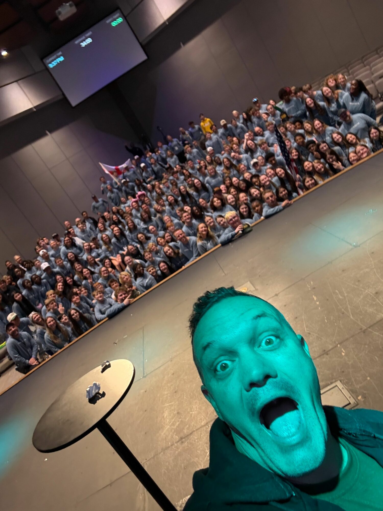 Person with surprised expression taking selfie in front of large crowd, with a lamp and wall in background.
