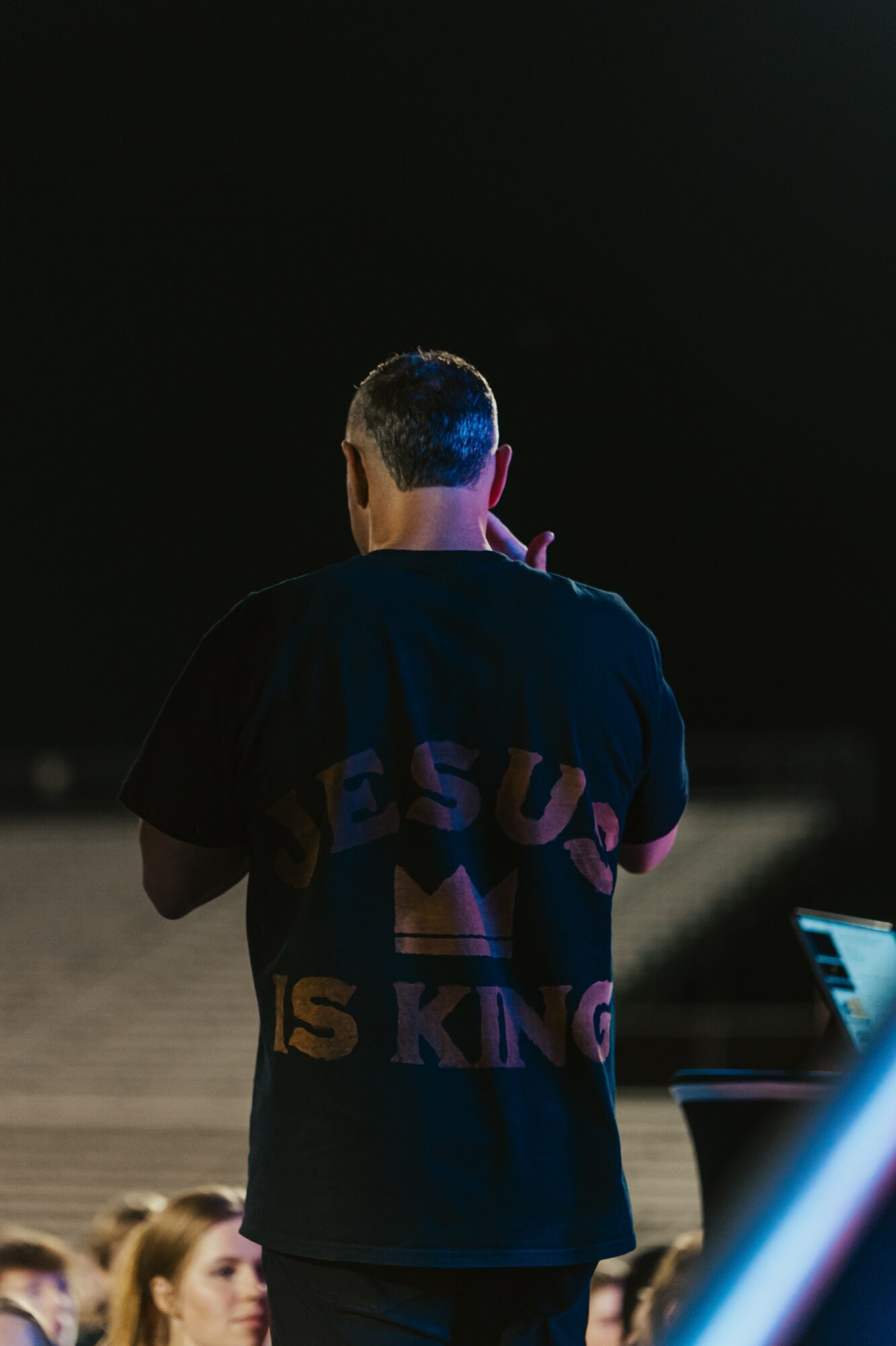 Person with short hair seen from behind, wearing a dark shirt with text, standing outdoors at night.