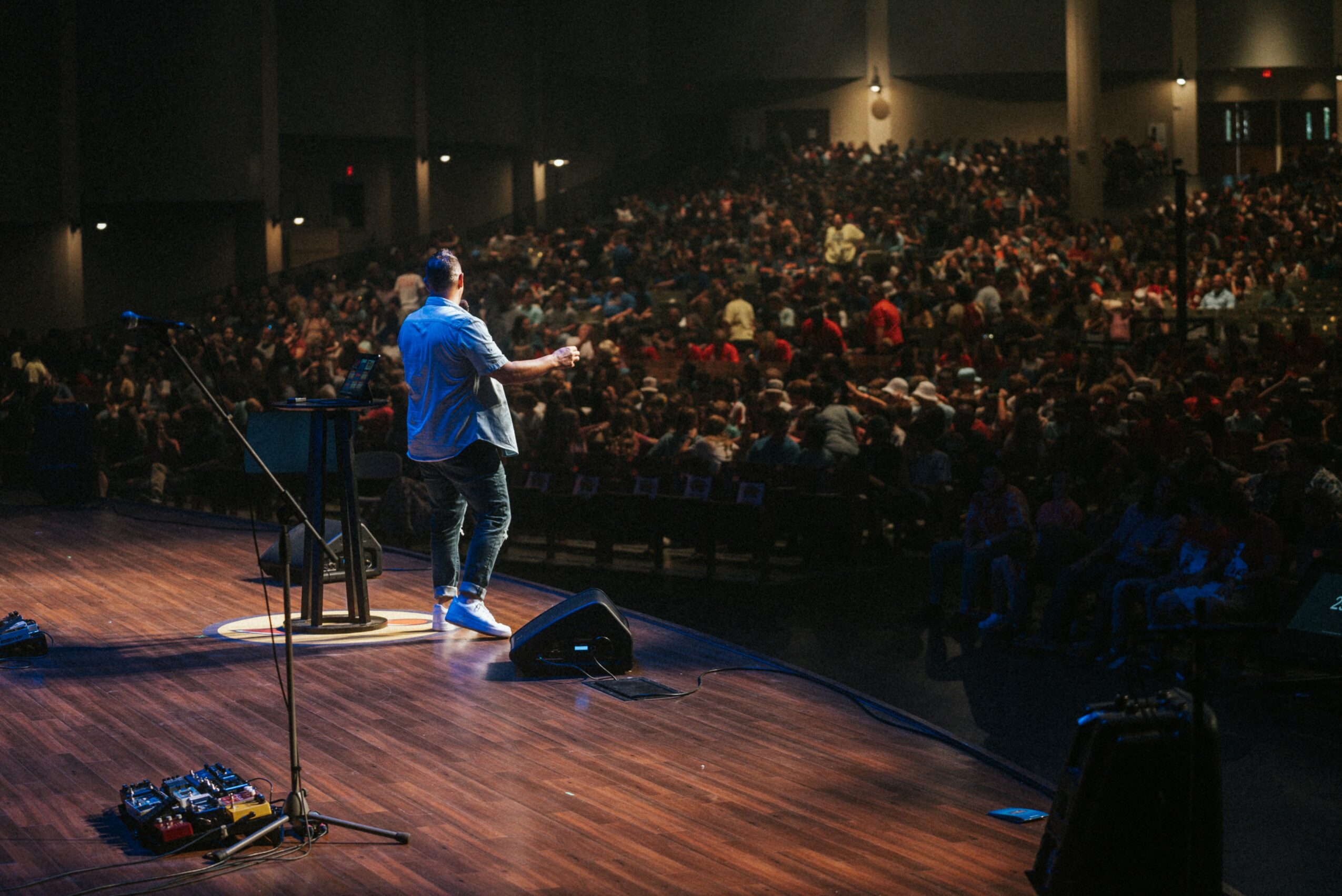 Performer on stage with guitar in front of large audience in auditorium, dim lighting, wooden stage floor.
