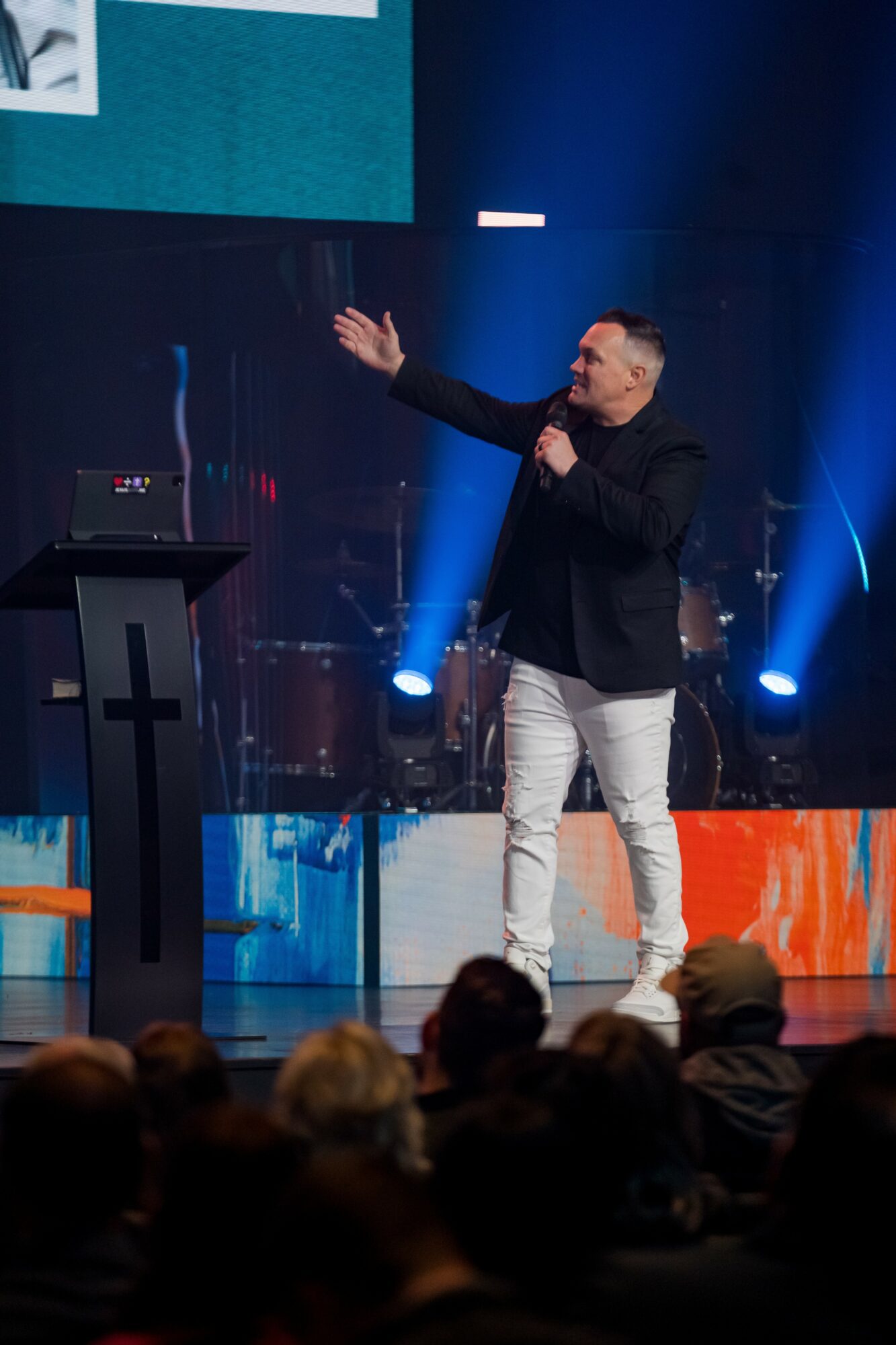 Man speaking on stage with a microphone, gesturing with his right hand, in front of a large screen and stage lights, audience visible.