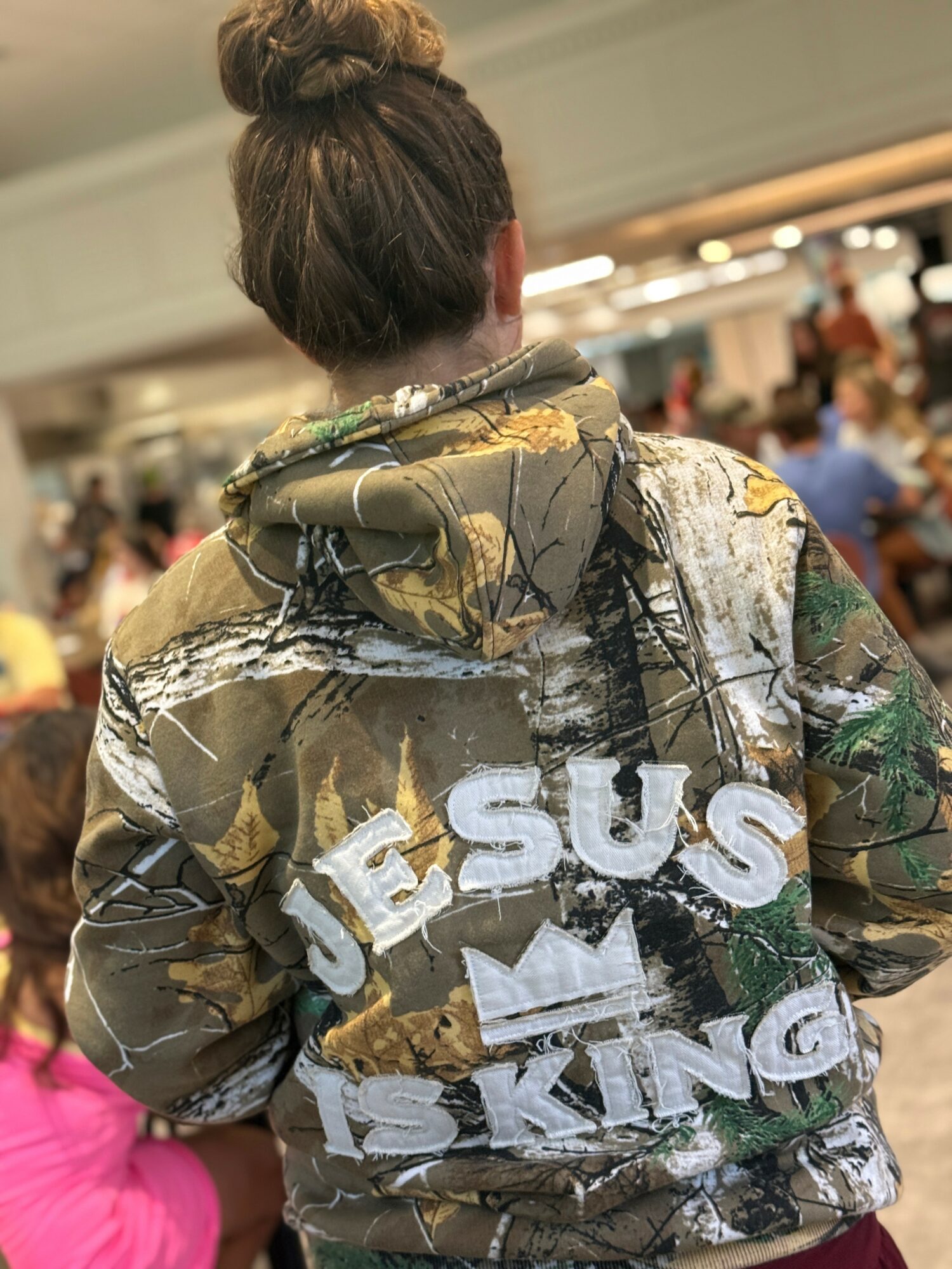 Back view of a person wearing a camouflage jacket with white text reading 'JESUS IS KING' in a crowded indoor setting.
