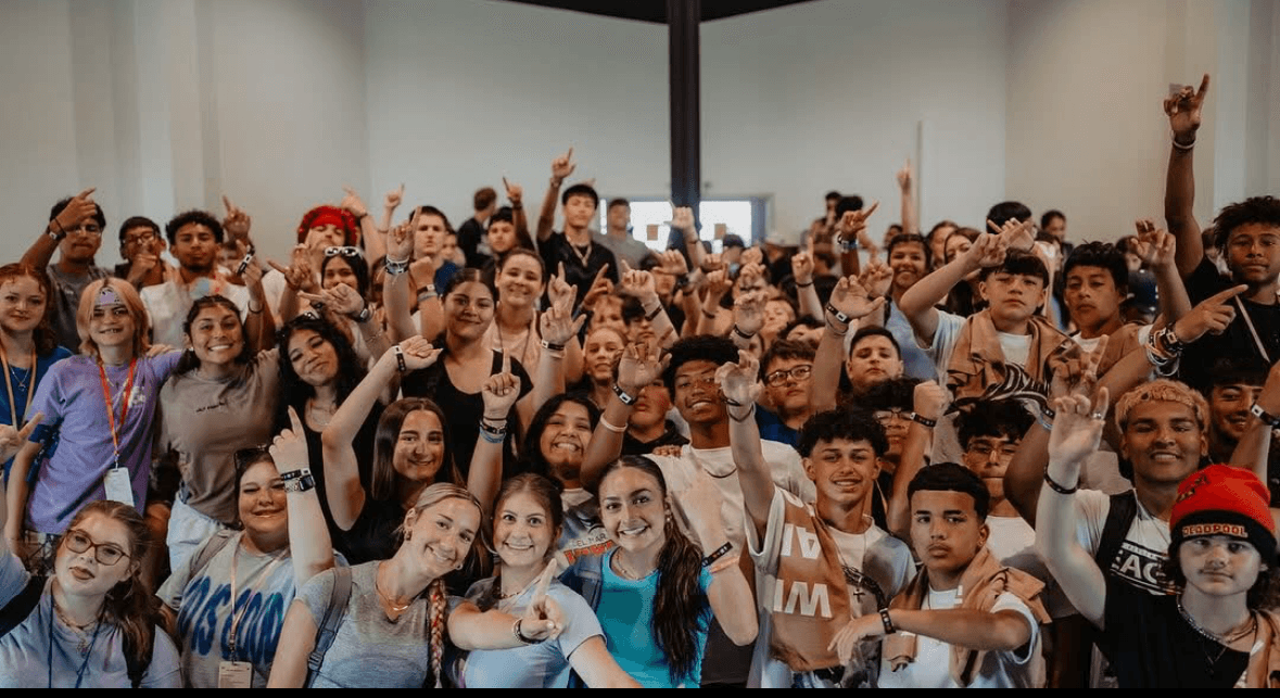 Large group of diverse young people gathered indoors, many raising hands and smiling.