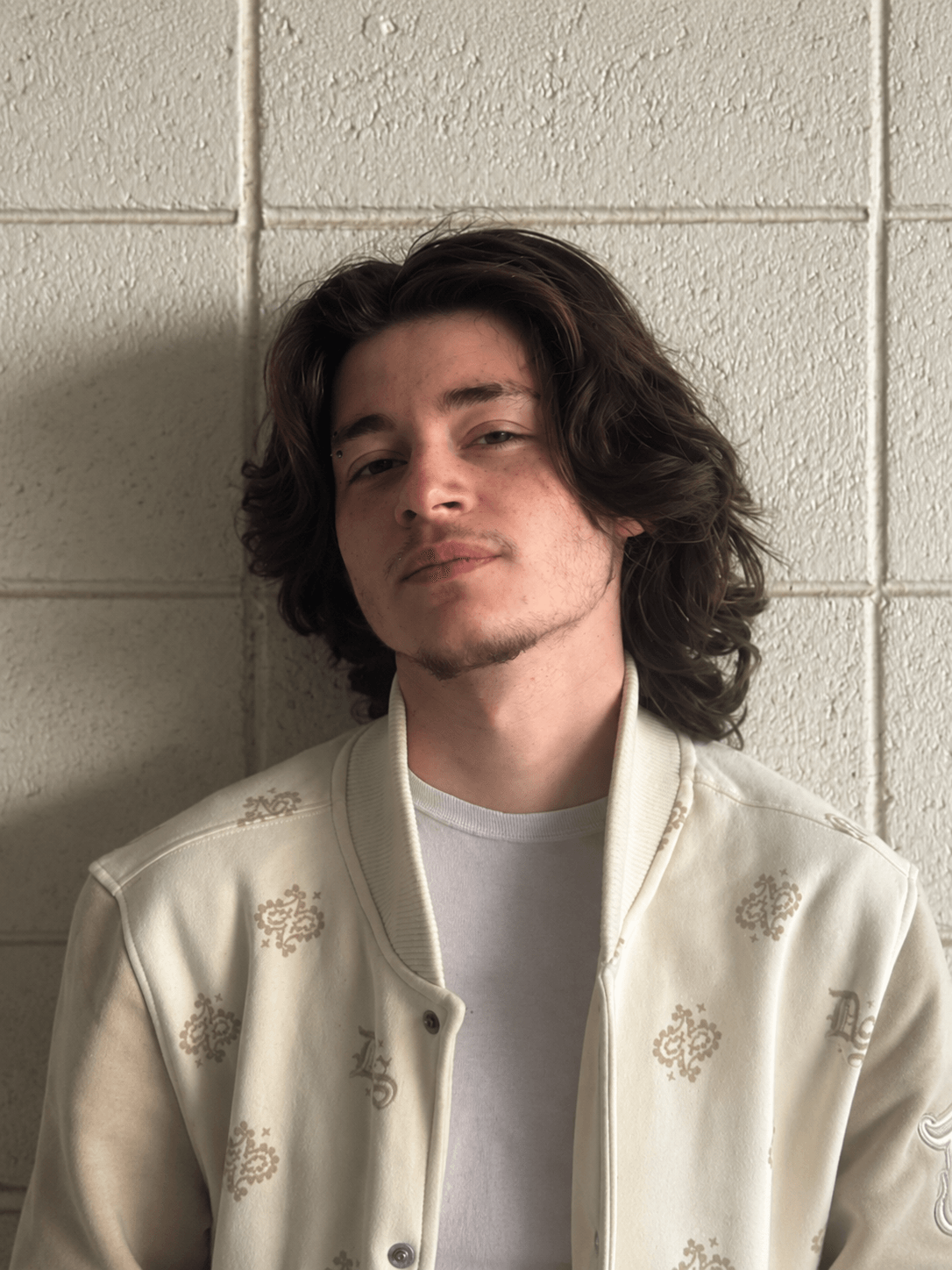 Young man with shoulder-length wavy hair and light facial hair, wearing a cream-colored jacket and white shirt, standing against a brick wall.