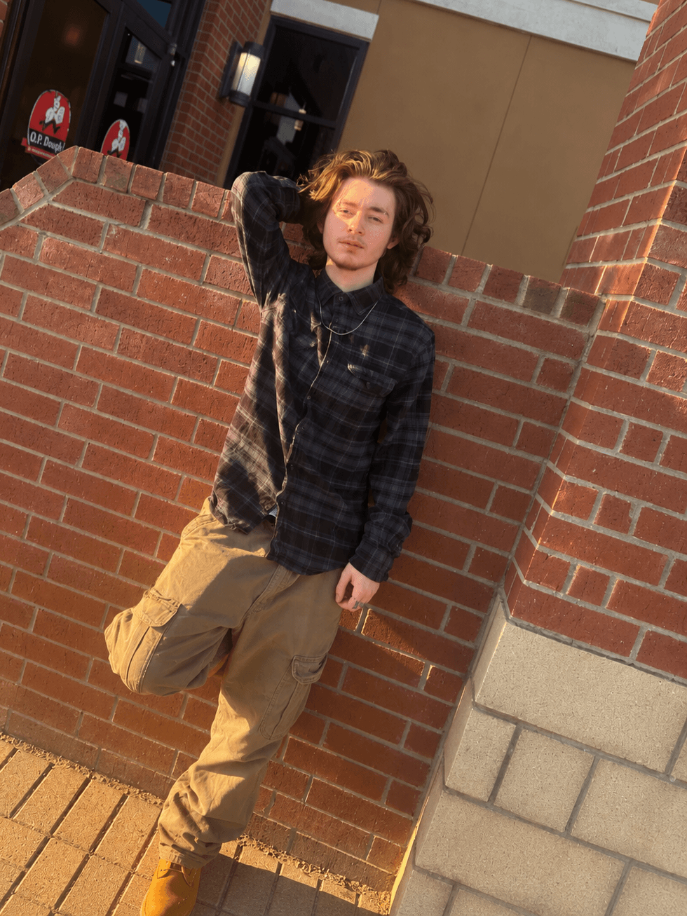 Young person with curly hair leaning against a brick wall outdoors, wearing a dark plaid jacket and beige pants.