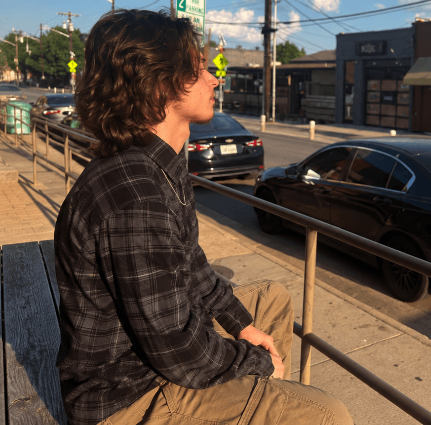 Person with curly hair sitting outdoors on a bench, wearing a plaid shirt, with cars and buildings in the background.