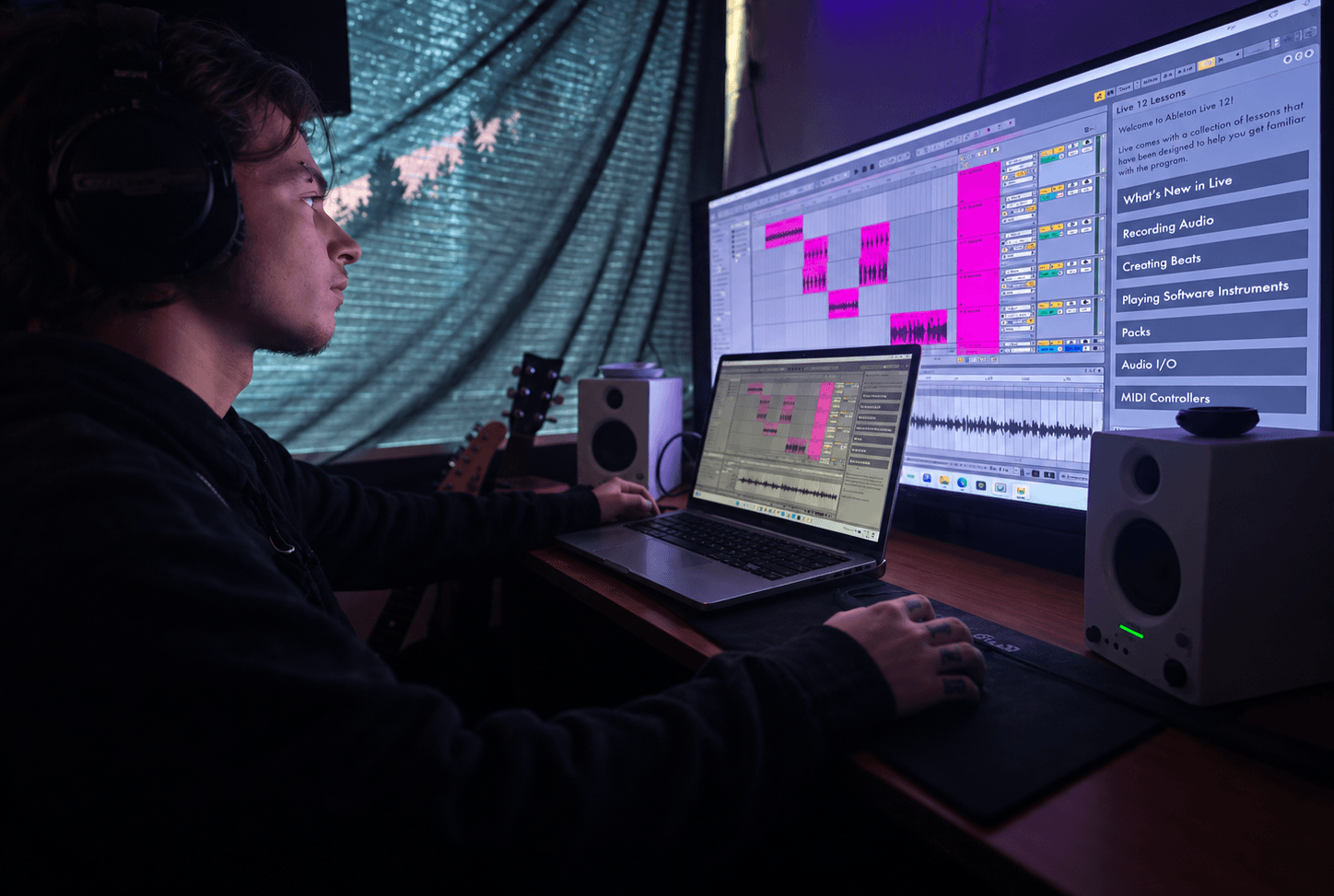 Person working on music production software with large monitor and laptop in a dimly lit room.