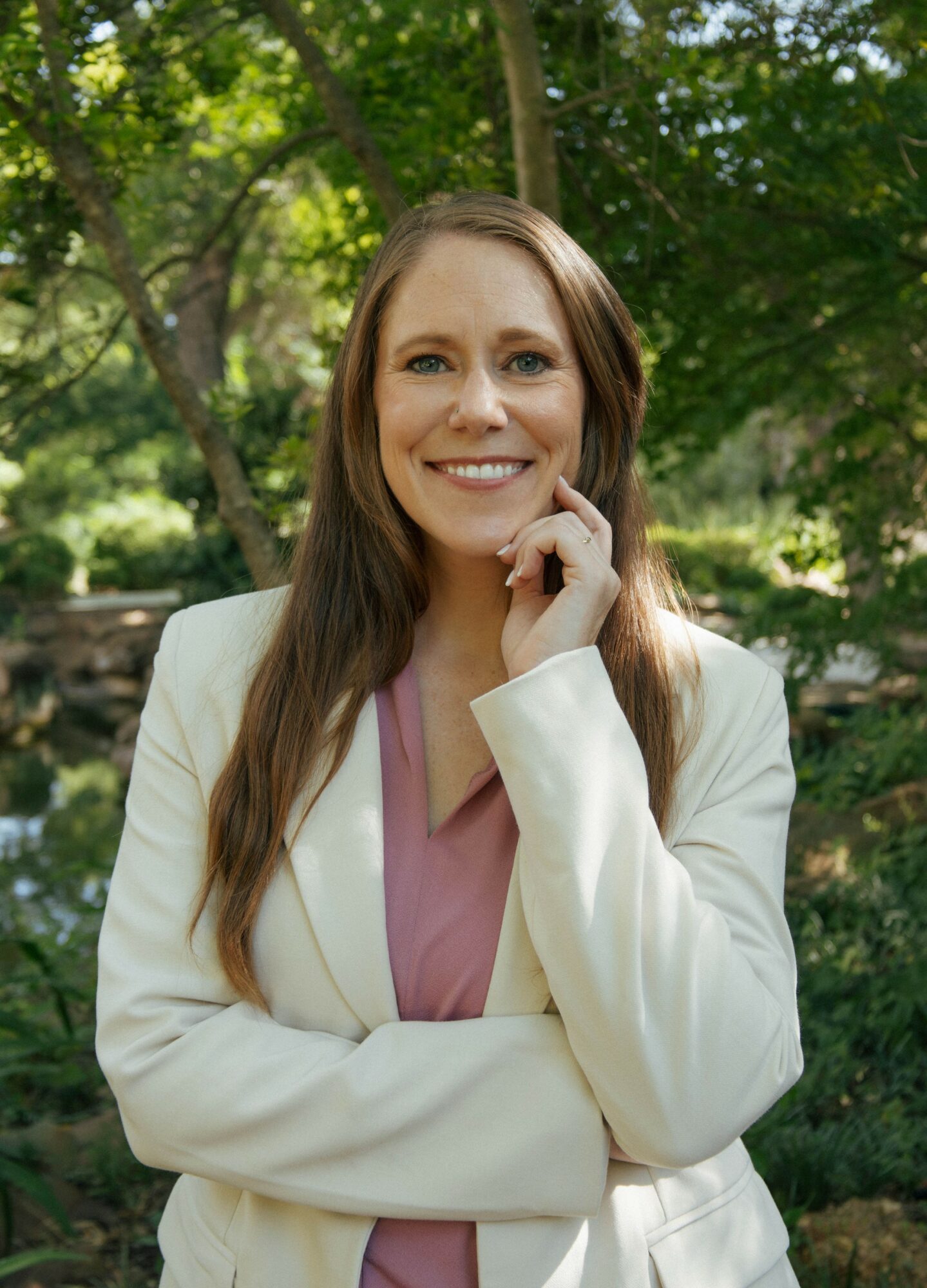 Woman with long brown hair smiling outdoors, wearing a white blazer and pink top, with trees in background.