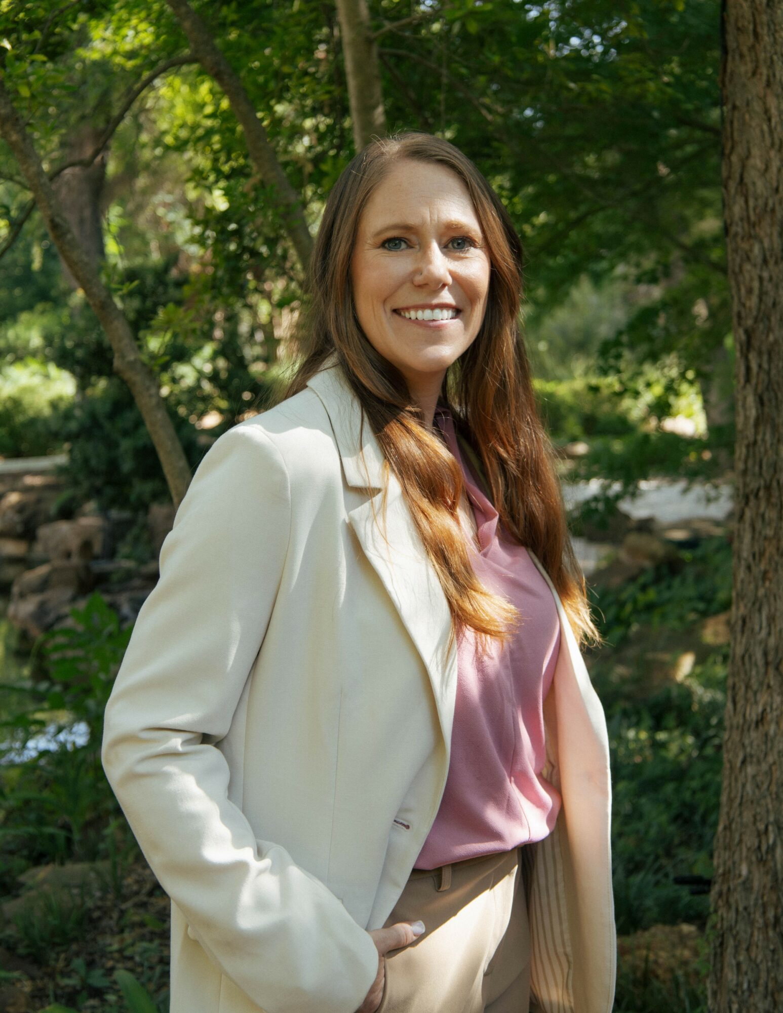 Woman with long hair smiling outdoors, wearing a light-colored blazer and pink top, surrounded by trees.
