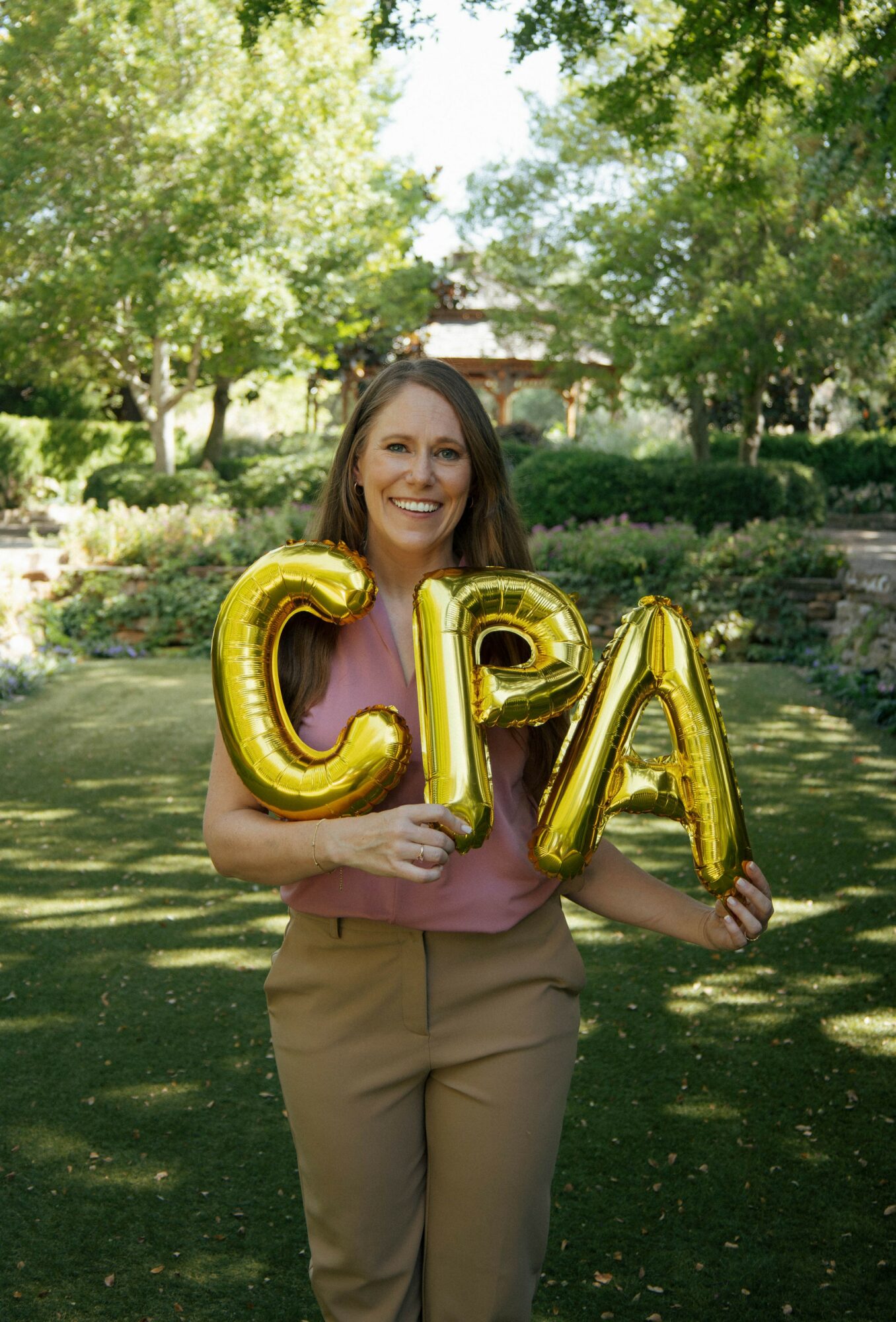 Smiling woman holding gold balloons spelling 'CPA' outdoors in a park with trees and sunlight.