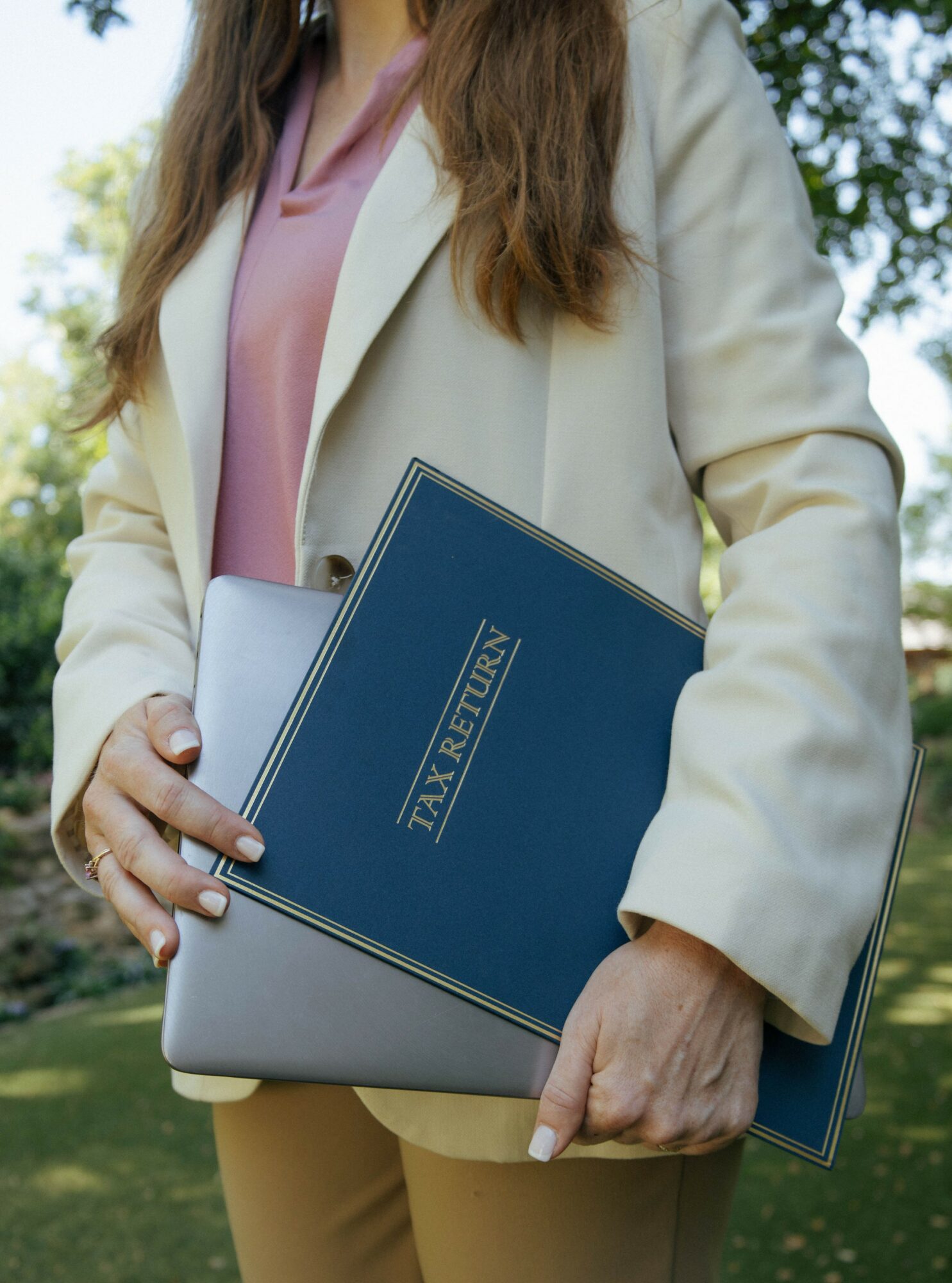 Person holding a diploma and a folder outdoors, wearing a beige blazer and pink top.