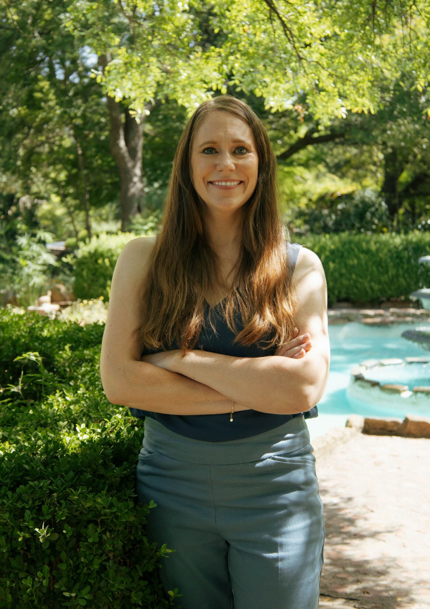 Woman with long hair smiling outdoors, arms crossed, in a park with trees and a pond in the background.