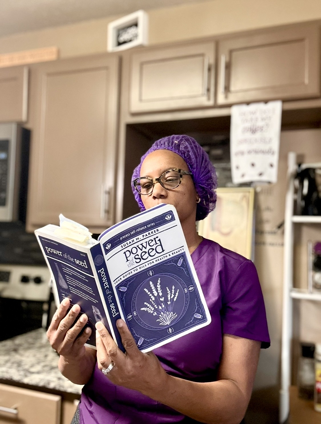 Woman wearing purple scrubs and a purple hair cover reading a book titled 'Poverty and Seeds' in a kitchen.