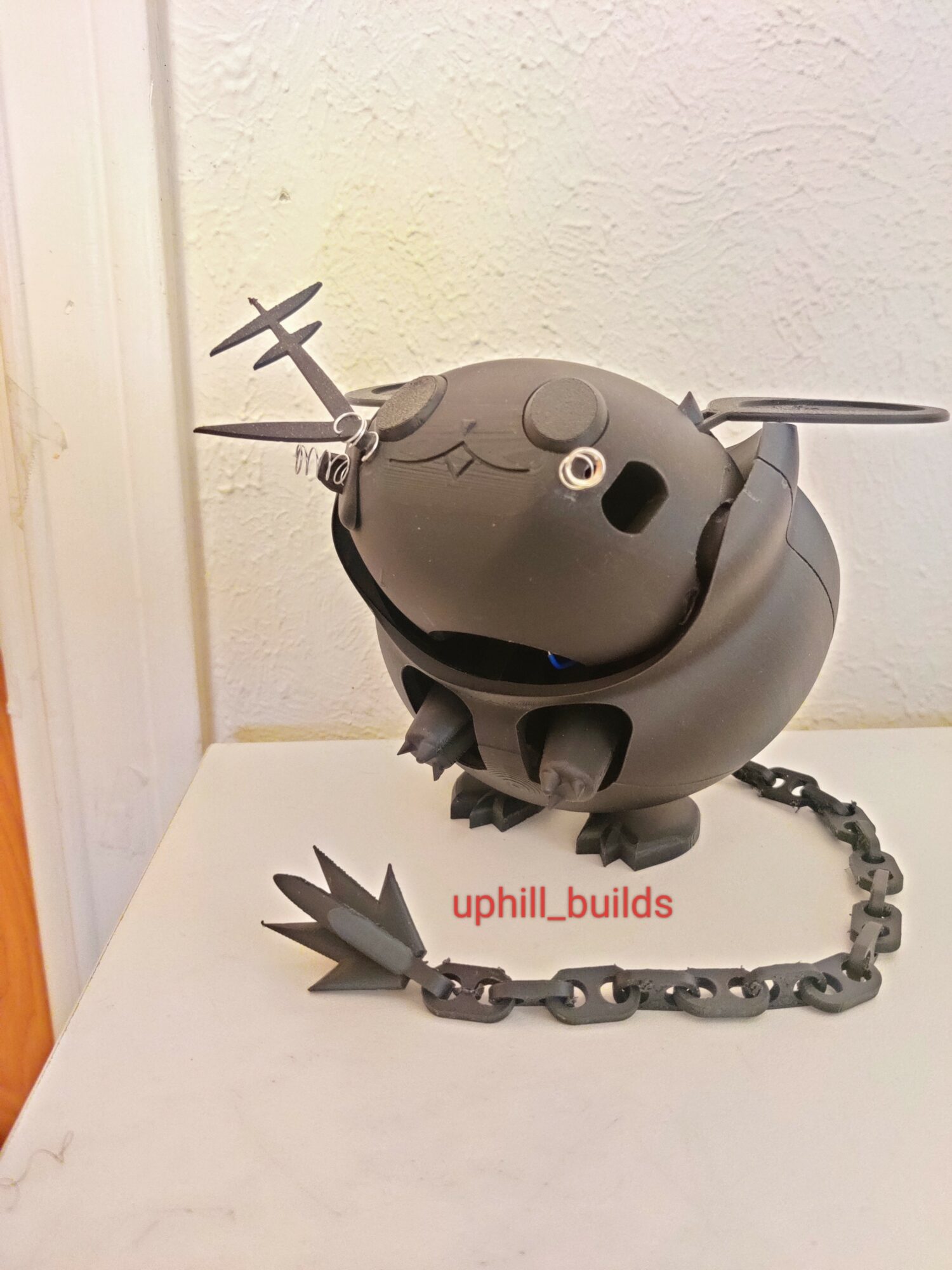 Black robot with a chain, antenna, and facial features, standing on a white surface against a textured wall.