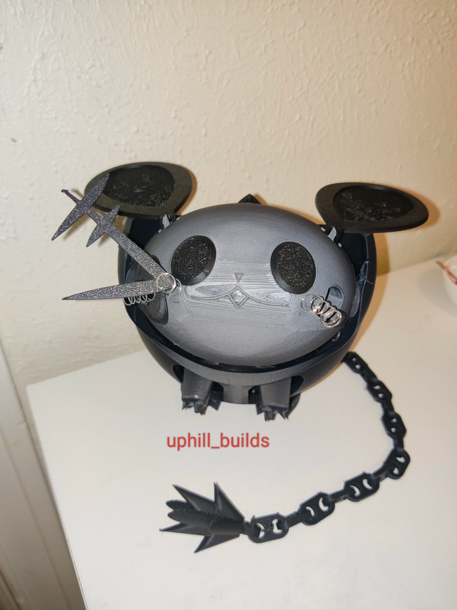 A robot with a round body, two black eyes, and two black antennae, with a chain attached to its side.