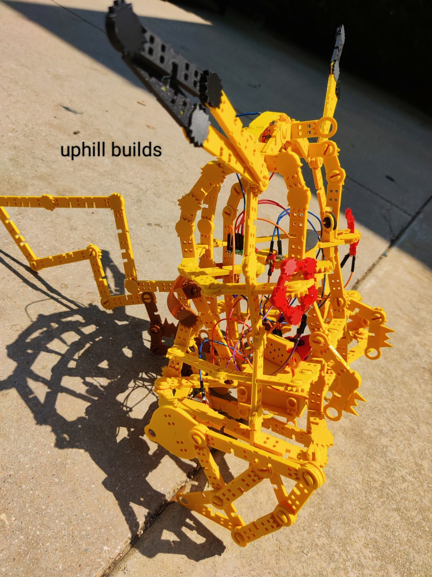 Yellow LEGO robot with mechanical arms and shadow on ground, sunlight casting shadows, labeled 'uphill builds'.