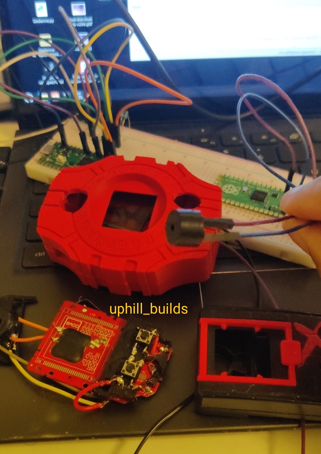 Red electronic device with a small screen, connected to wires, on a workspace with circuit boards and tools.