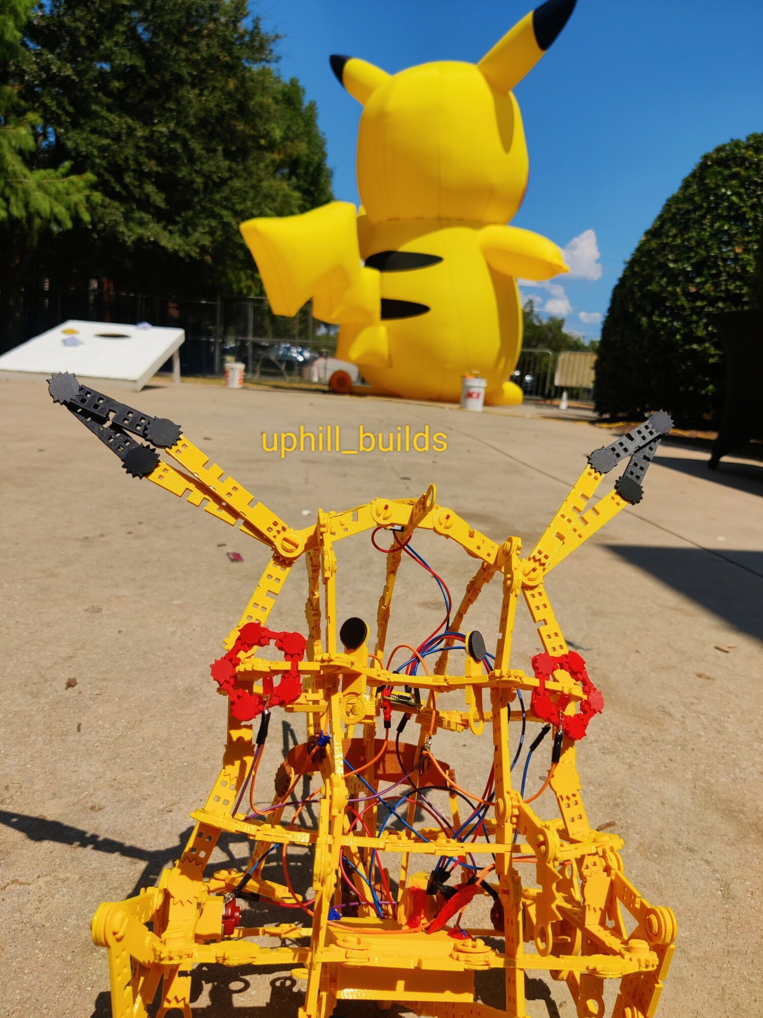 Large Pikachu sculpture in park with trees, blue sky, and a yellow roller coaster in front, made of LEGO bricks.