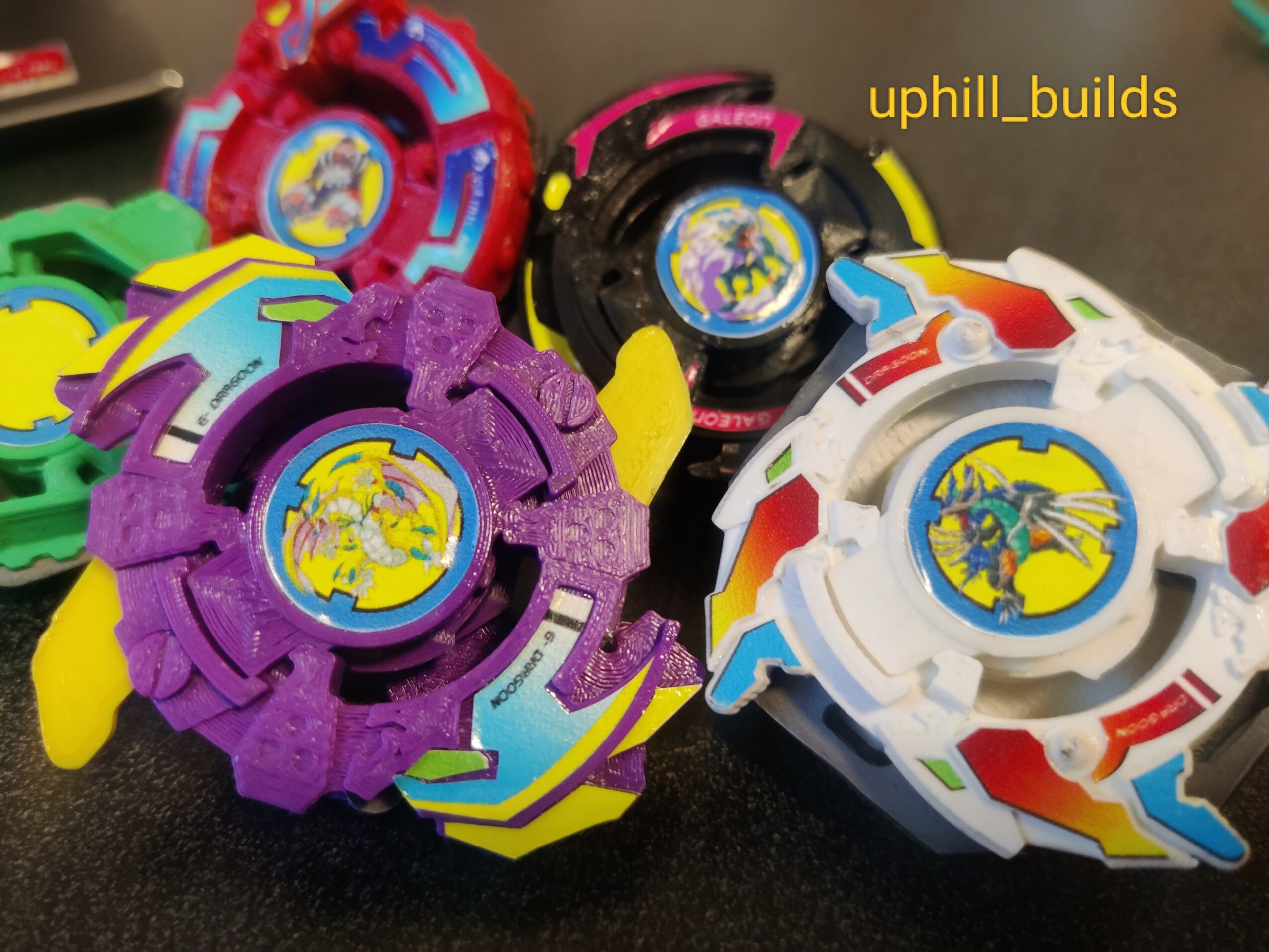 Four colorful Beyblade toys with dragon logos, in purple, white, black, and green, on a dark surface.