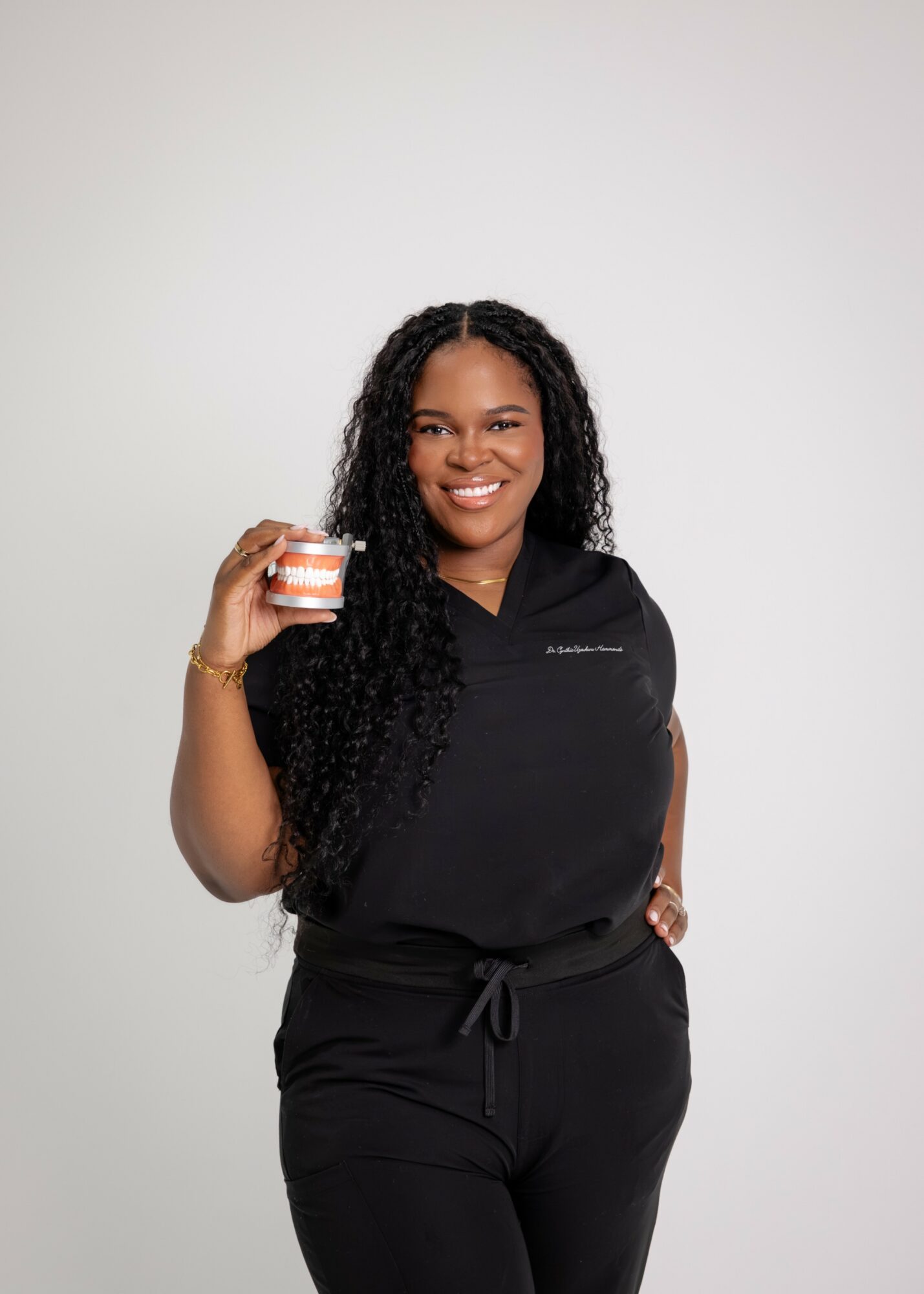 Smiling woman with long curly hair holding a small container, wearing a black outfit, standing against a plain background.