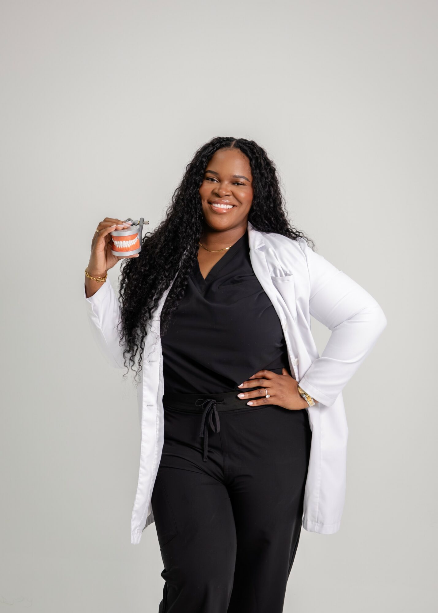Woman with long curly hair smiling, holding a small container, wearing a black outfit and white coat, standing against a plain background.