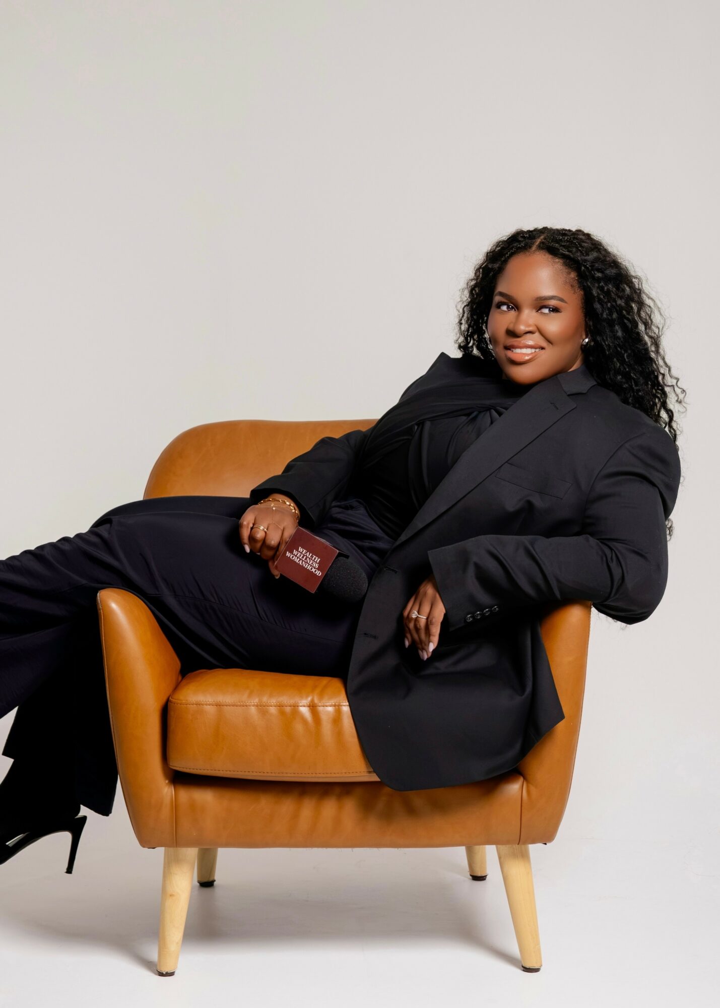 Woman with curly hair sitting on a tan armchair, smiling, holding a book, wearing a black blazer and pants, against a plain background.