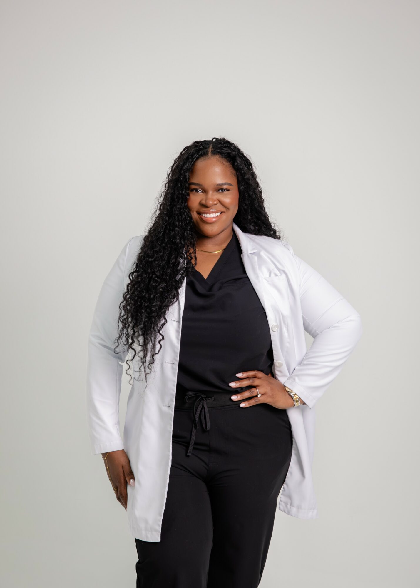 Woman with long curly hair smiling, wearing a white coat over black clothing, standing with hand on hip against a plain background.