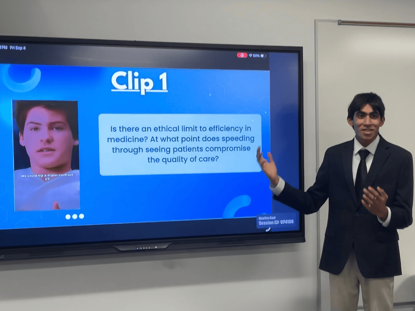 Young man in a suit presenting a slide on a large screen in a classroom, with a whiteboard nearby.