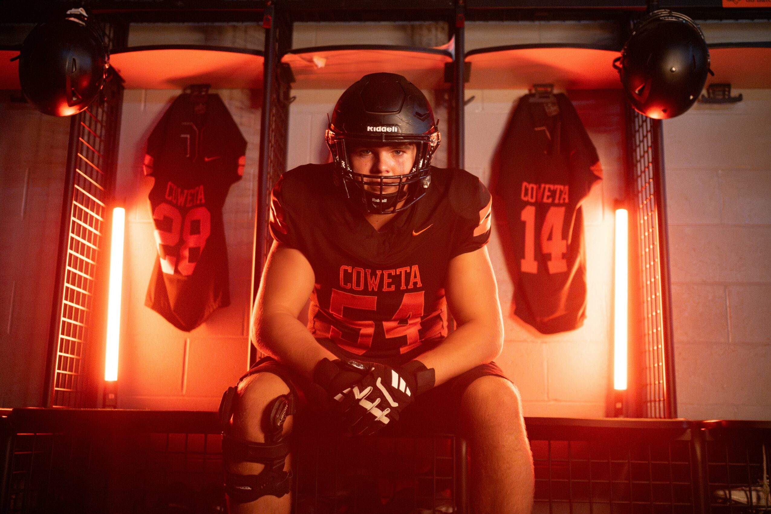 Football player in helmet and uniform crouching in a locker room with jerseys and red lighting behind.