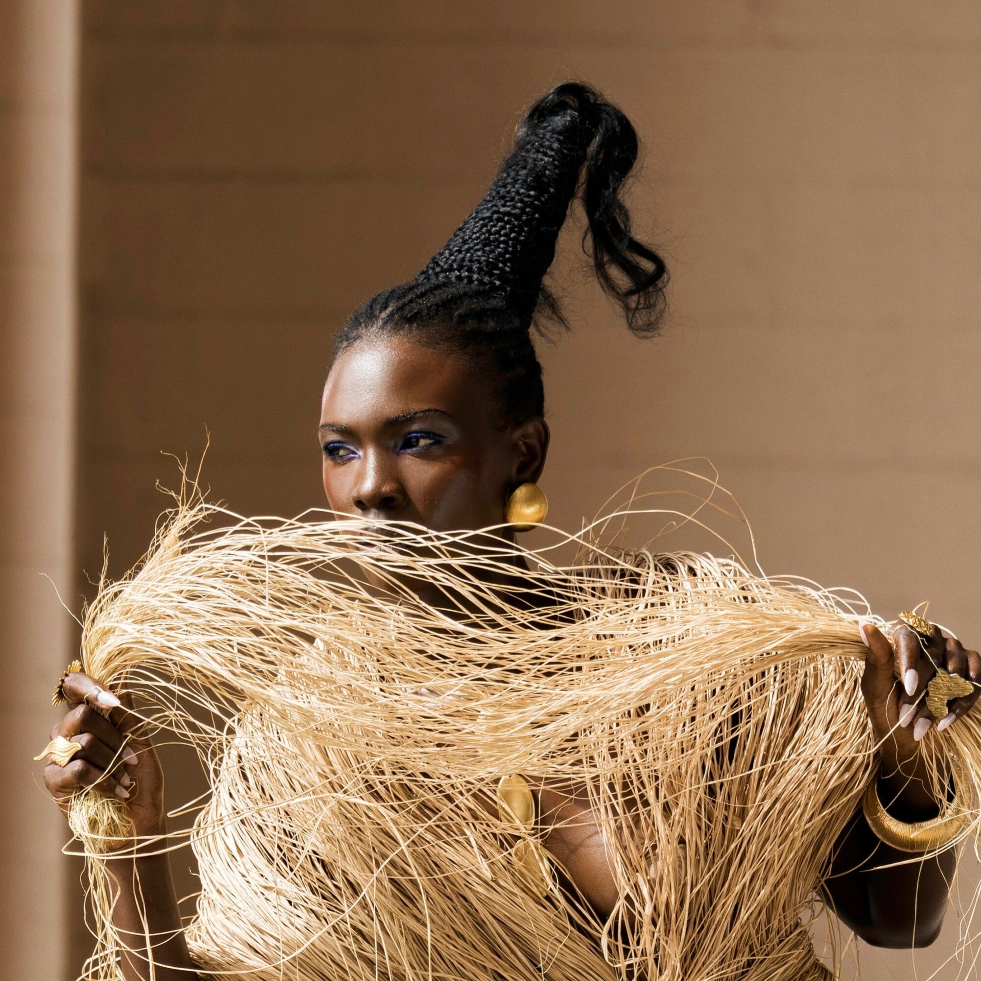 Woman with elaborate hairstyle holding a large, textured, golden accessory in front of her face, against a plain background.