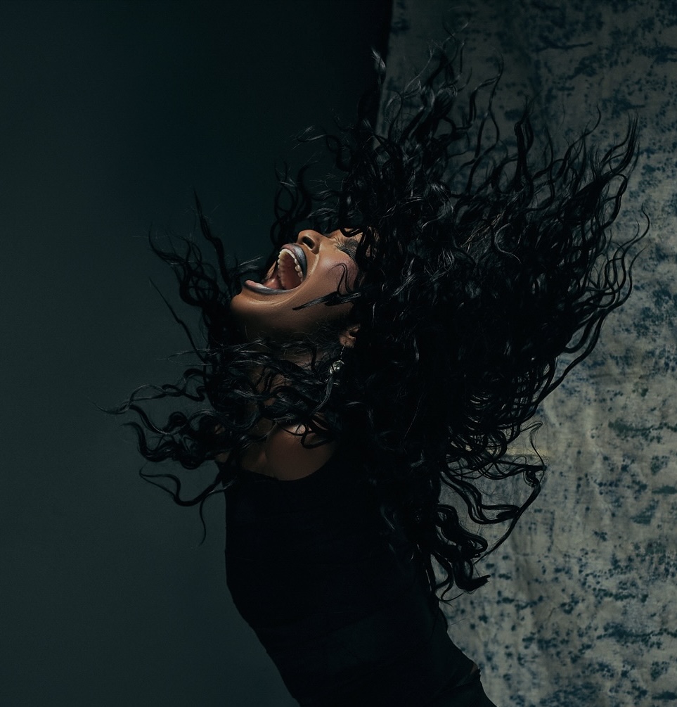 Person with long, curly hair flipping head back, mouth open, dark background, textured wall, expressive pose.