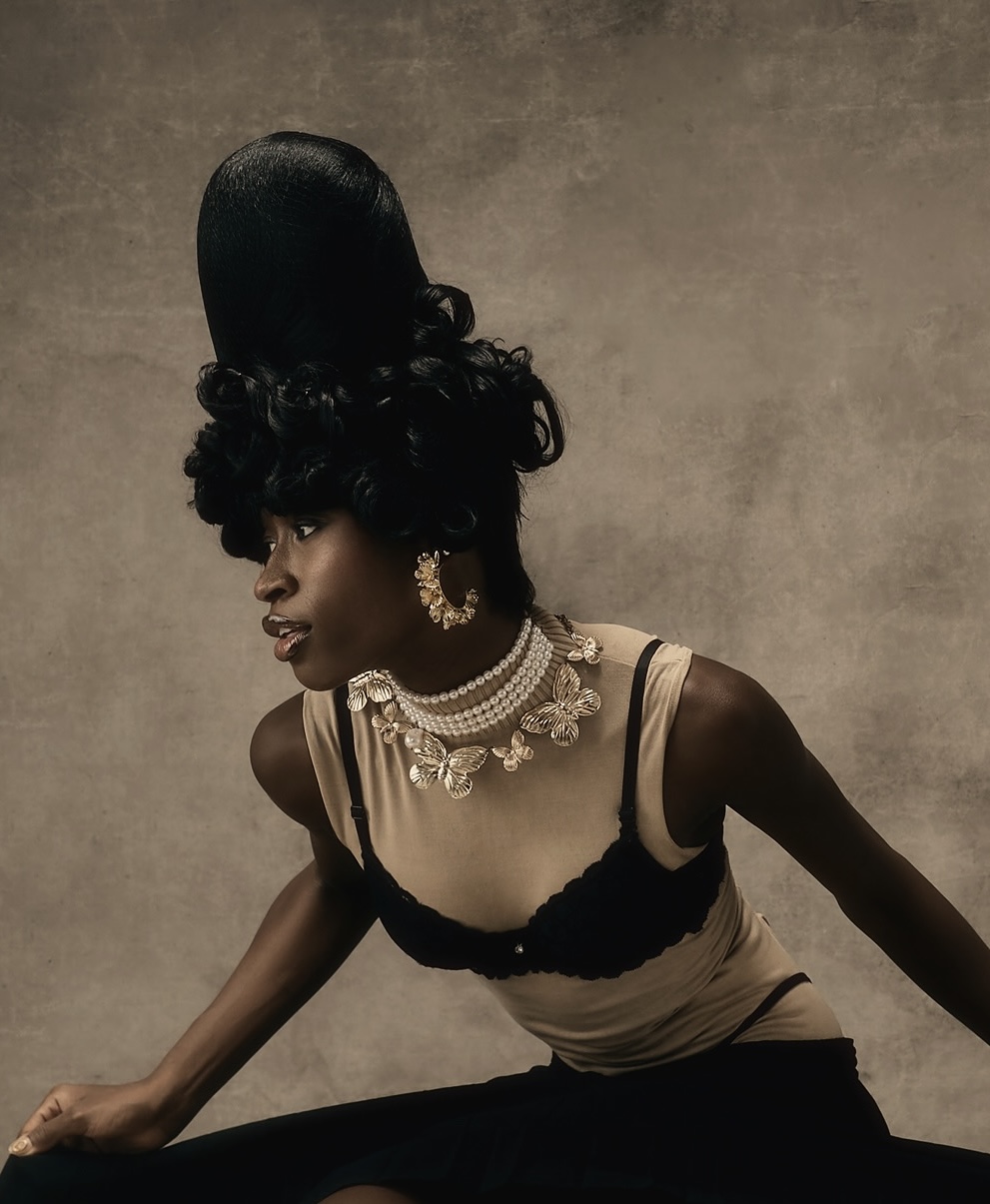 Woman with elaborate hairstyle and jewelry, wearing a black and beige top, posed against a plain background.
