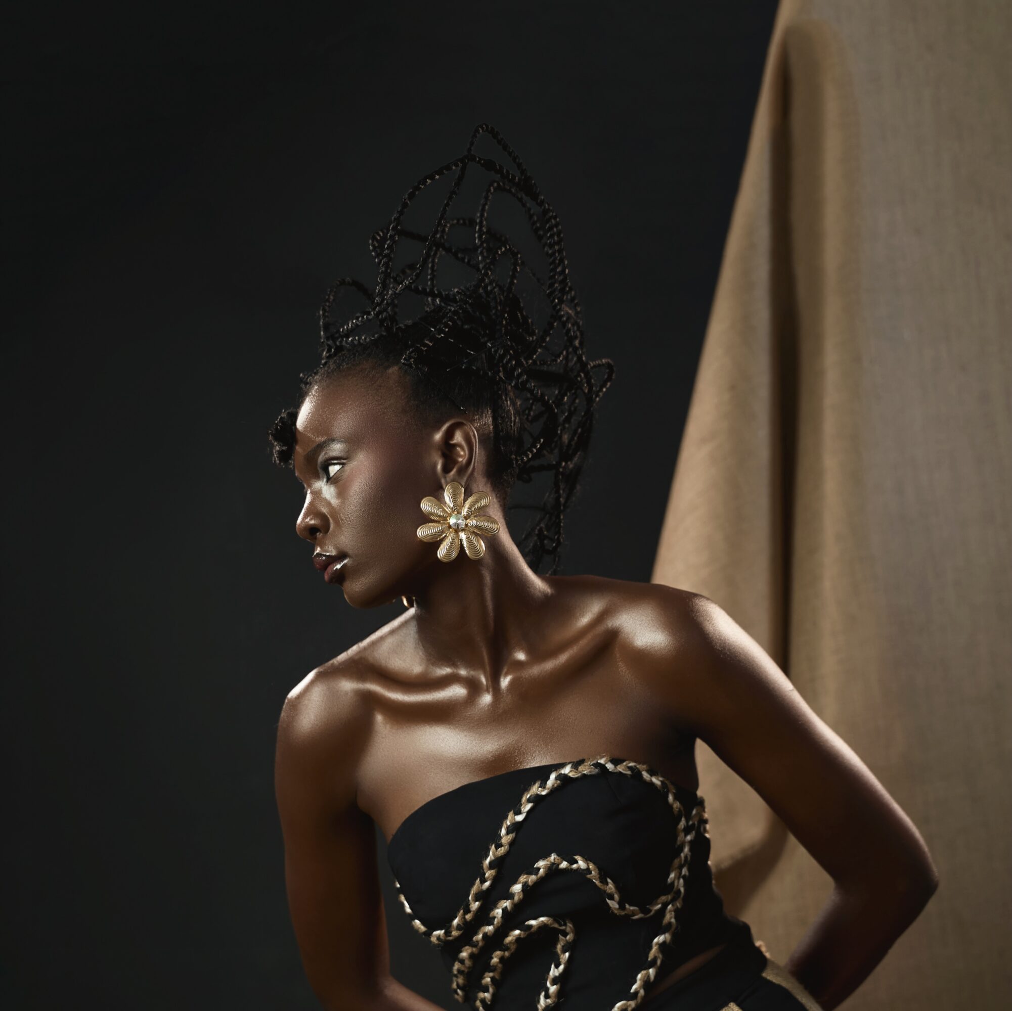 A woman with styled hair and large earrings poses against a dark background with a beige drape.