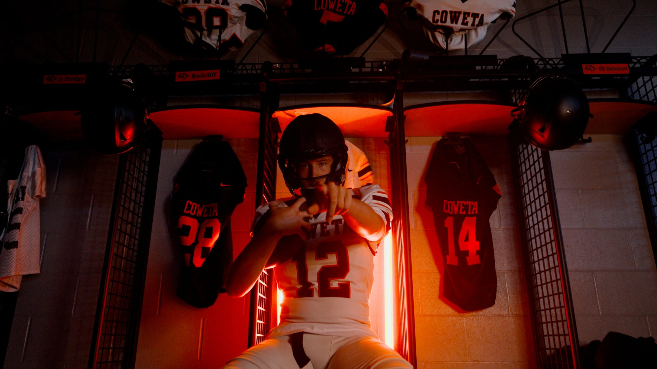 Football player sitting in locker room with jerseys and equipment, illuminated by red lighting, holding a helmet.